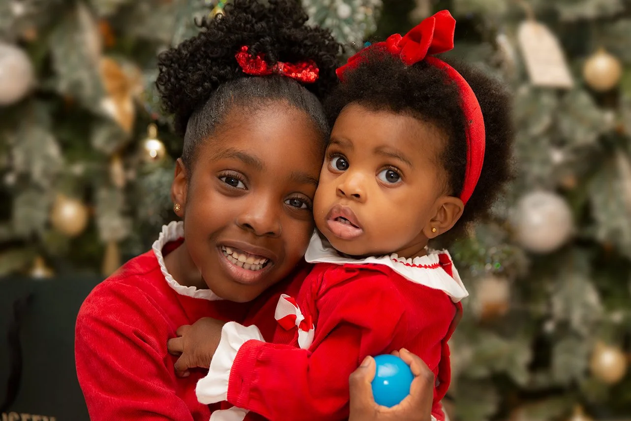 Two young girls dressed in red holiday outfits hugging each other in front of a decorated Christmas tree.