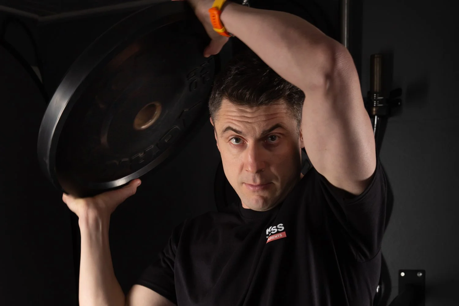 A man holding a large black record on his shoulders, looking directly at the camera, in a dark room.