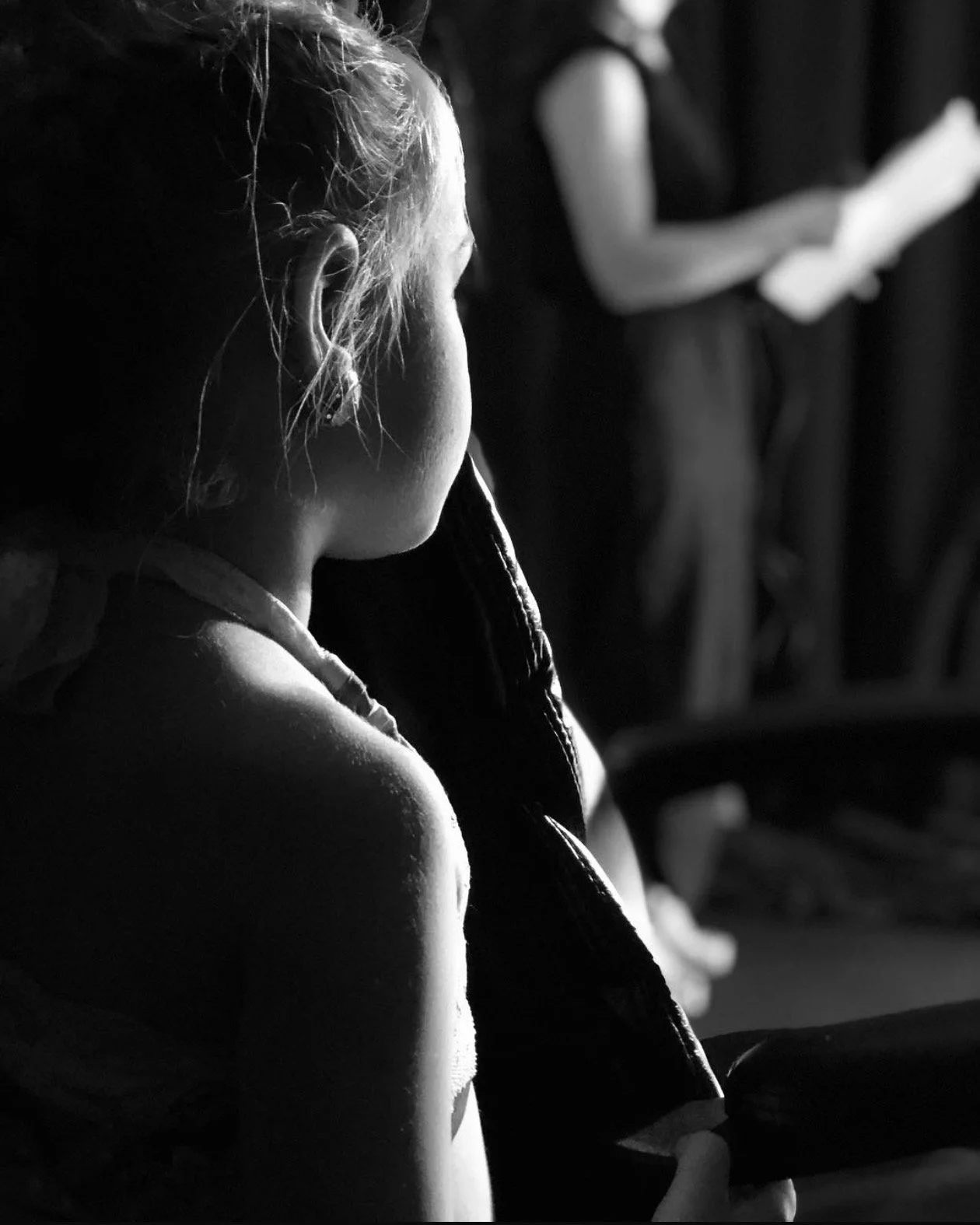 A young girl with her hair tied back, seen in profile, holding a book or notebook, while another person stands in the background reading from a paper.
