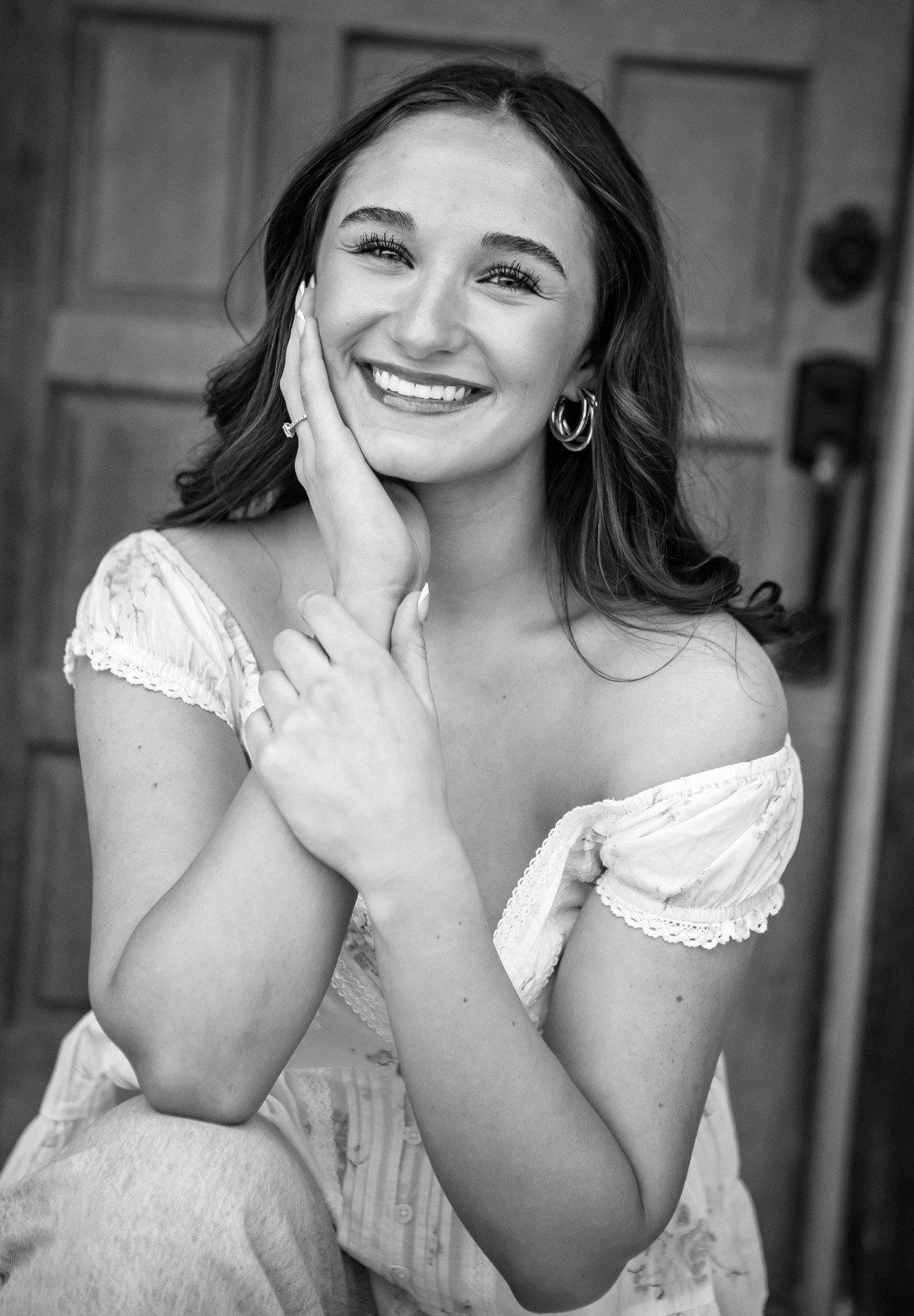 A smiling woman with wavy hair, wearing hoop earrings and a light-colored off-shoulder blouse, posing with her hand on her face in front of a wooden background.