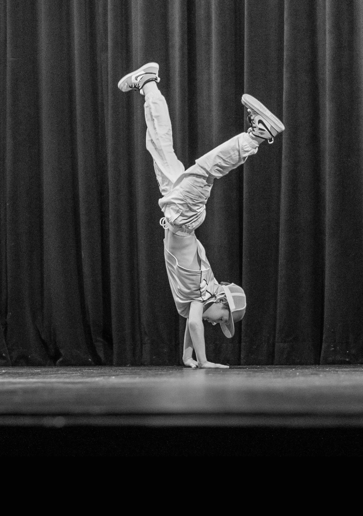 A young person in casual clothing, including a cap, performing a handstand on a stage with a dark curtain behind them.