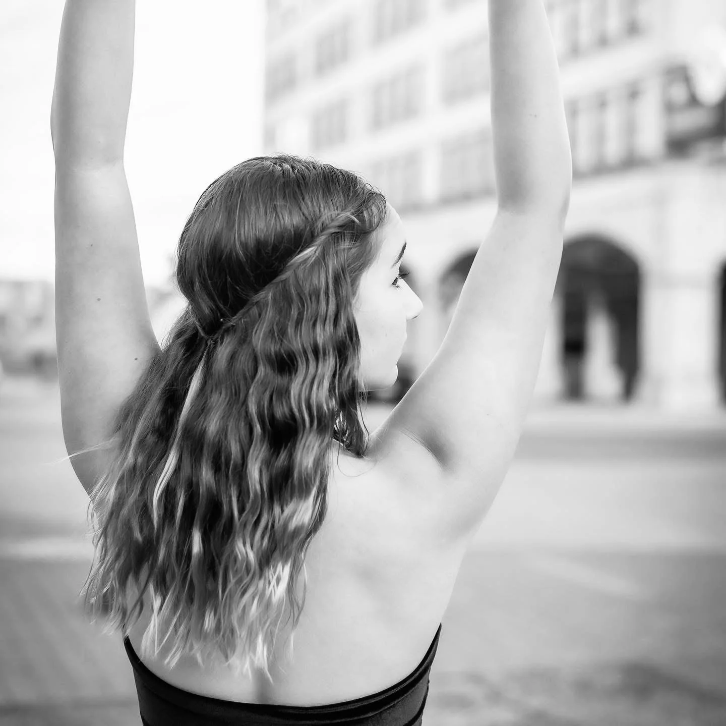 A young woman with wavy hair stretching her arms up outdoors, with blurred city buildings in the background.