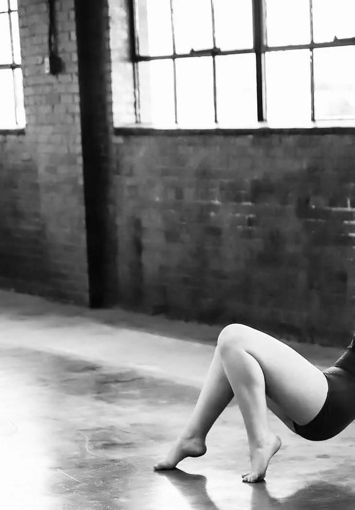 Close-up of a person doing a ballet pose on a polished floor inside an industrial-style studio with brick walls and large windows.
