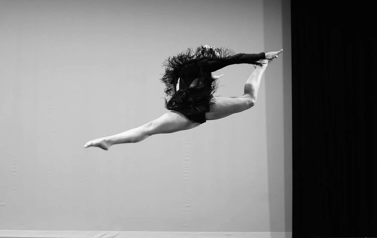 Black and white photo of a female dancer, mid-air, in a split leap, with her long hair flowing behind her, against a plain background.