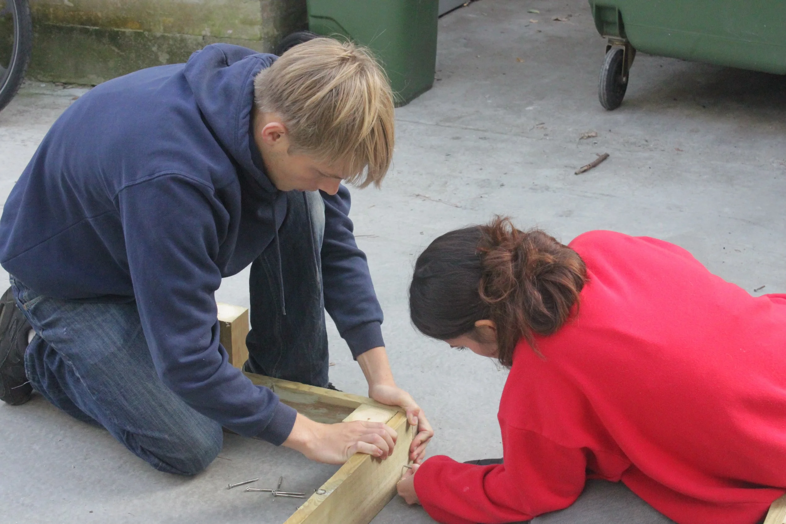 To personer, en mand i blå sweatshirt og en kvinde i rød sweatshirt, arbejder sammen på et træstykke, de sidder på knæ på græsplæne og fastgør det.