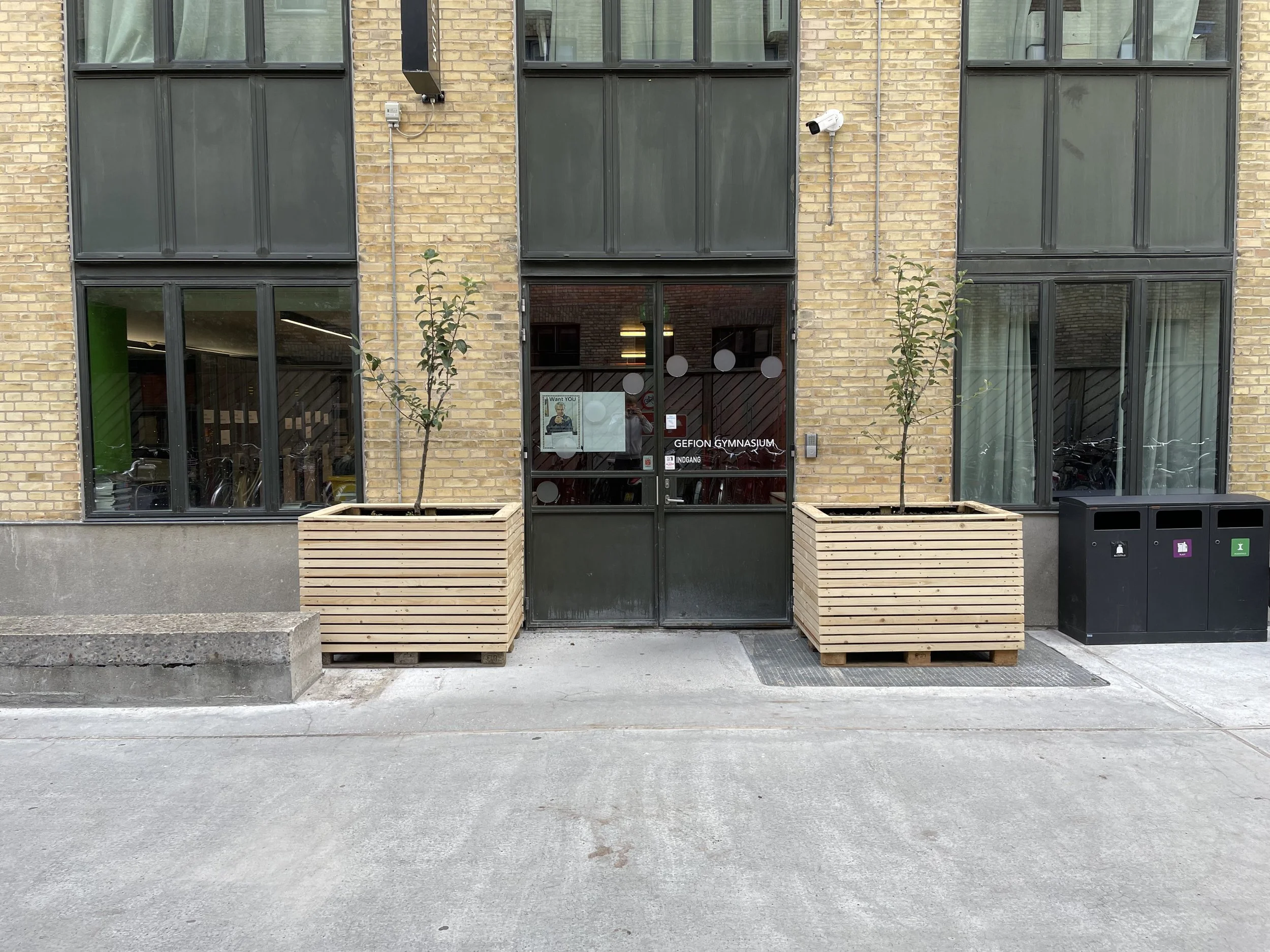 The image shows the entrance to a gymnasium building with large windows and a door. There are two wooden planters with small trees on either side of the door, a security camera mounted above, and three trash bins on the right side.