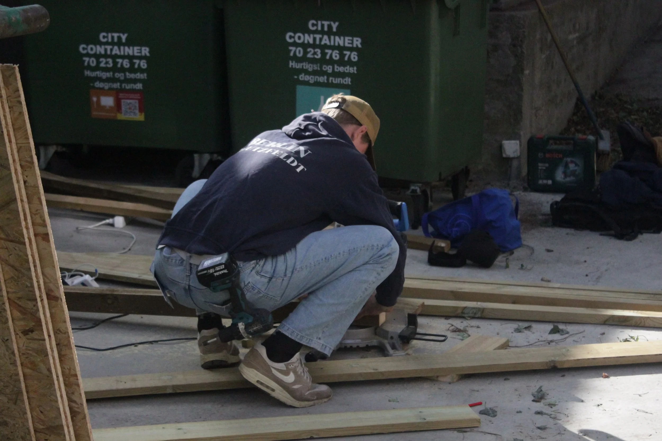 En person arbejder med træ på et udendørs arbejdsområde med skraldespande og værktøj i baggrunden.