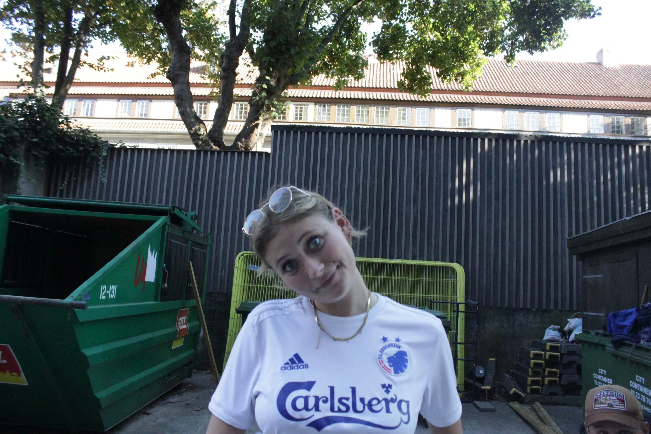 En kvinde med blond hår, solbriller på hovedet, iført en hvid t-shirt med logo og teksten 'Carlsberg', poserer udendørs foran en sort stålplade, containere og træer i baggrunden.