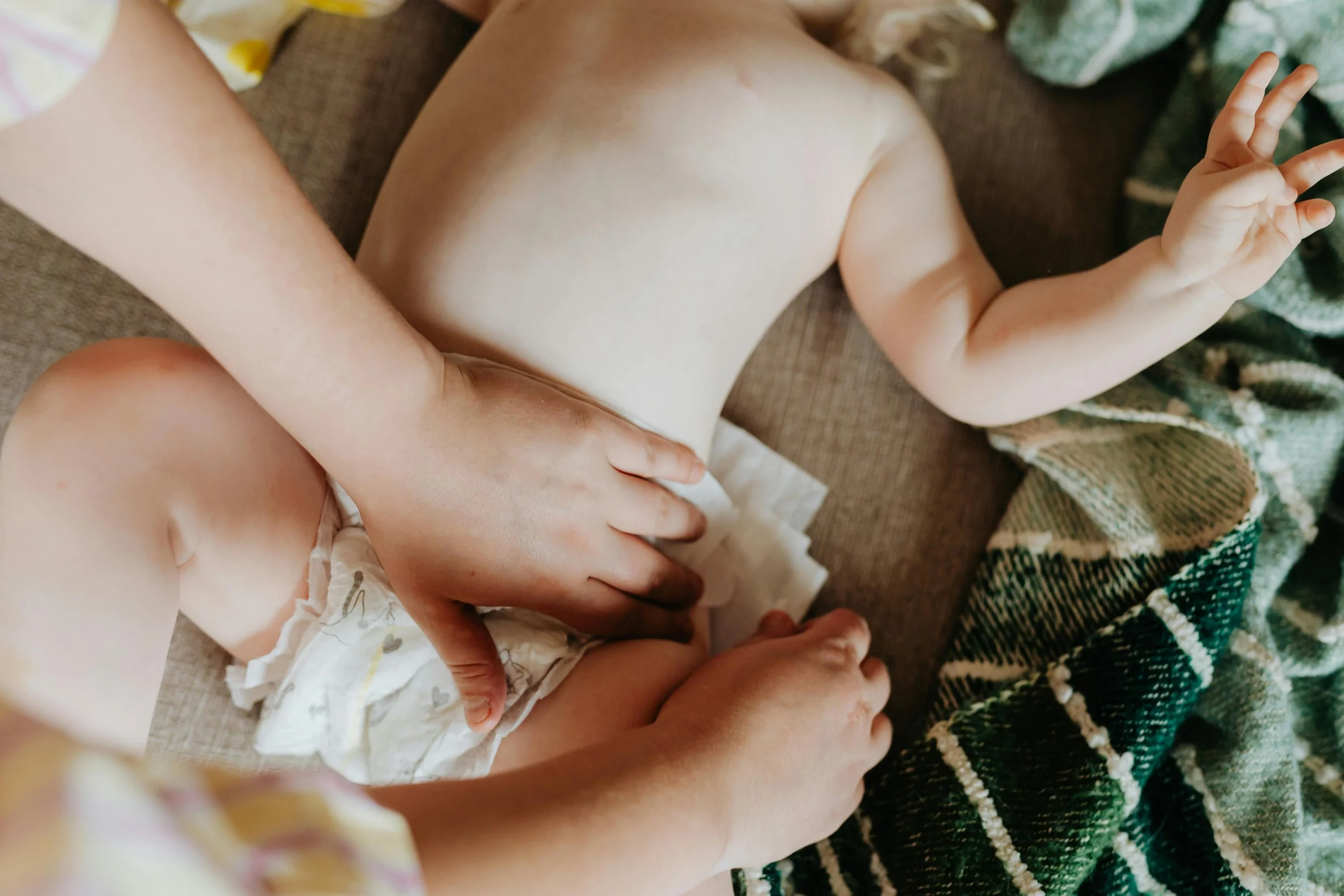 A baby lying on a couch with an adult holding and supporting them. The baby's torso and arms are visible, with the adult's hand on the baby's stomach. The baby is wearing a diaper and has their hand raised, making a peace sign with their fingers. There is a blanket with a plaid pattern nearby.