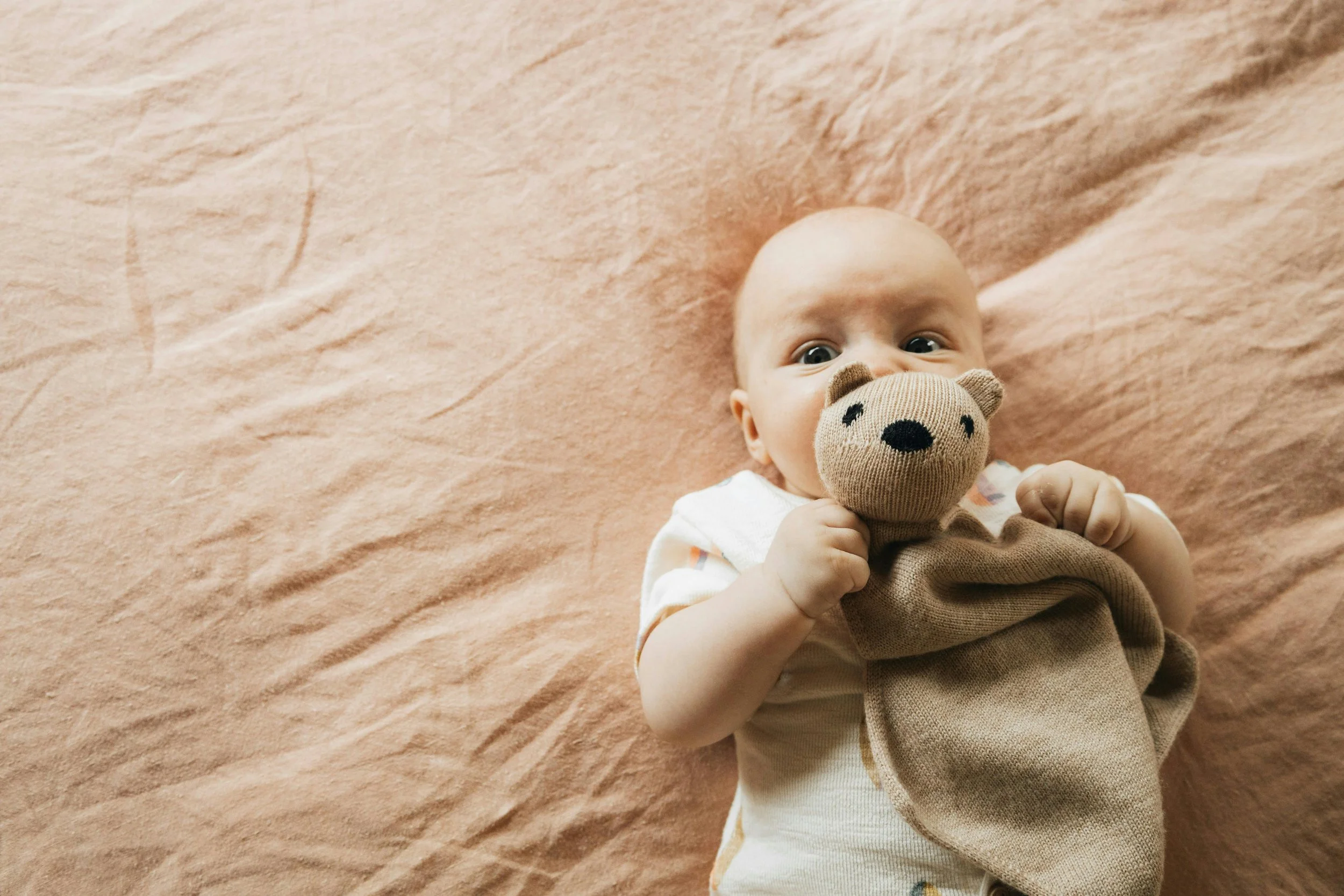 A baby lying on a soft, pink blanket, holding a stuffed animal sock monkey close to their face.