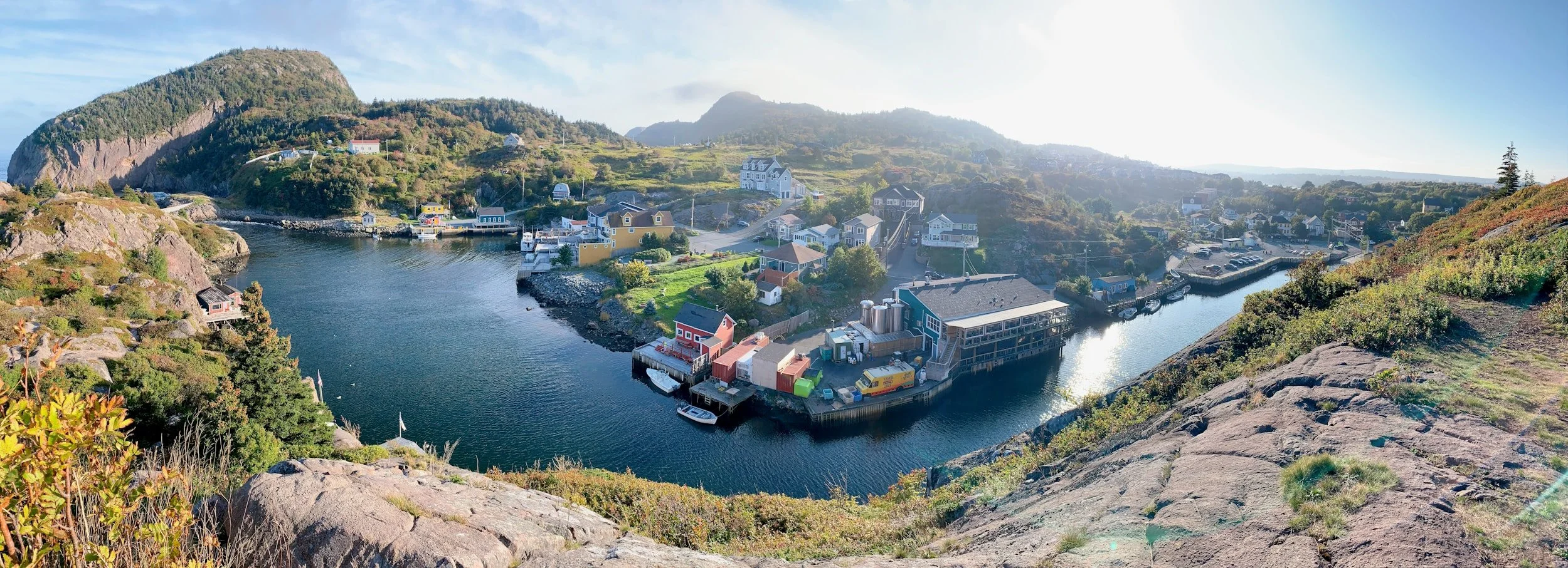 A scenic view of a harbor town with colorful houses, boats, and a rocky hillside in the background, taken from an elevated vantage point.