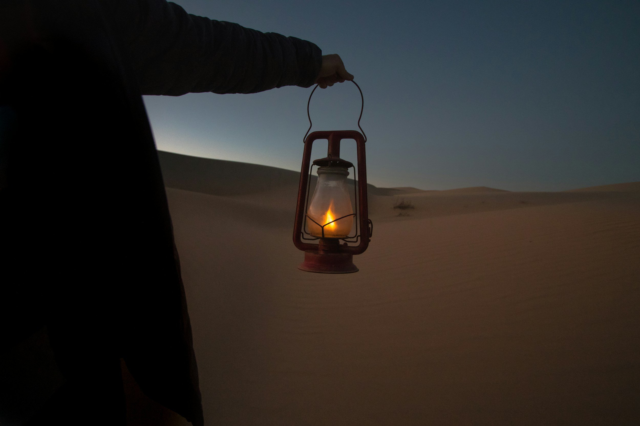 A person holding a lantern with a lit candle inside, standing on a sand dune in a desert at dusk or dawn.