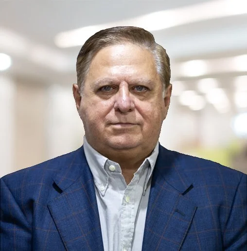 A middle-aged man with light skin, short brown hair, and a serious expression, wearing a light-colored shirt and a blue plaid blazer, standing in an indoor setting with blurred background lights.