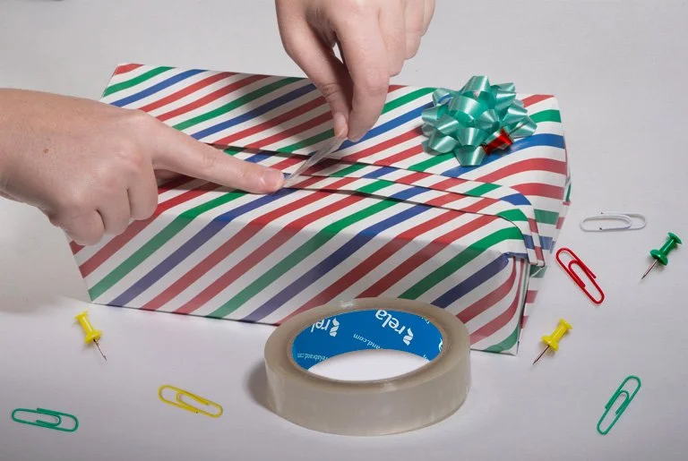Hands wrapping a gift with striped red, green, blue, and white paper, using clear tape and decorated with a shiny green bow. Scattered around are colorful paper clips, a roll of clear tape, a yellow push pin, a red paper clip, and a green push pin on a white surface.
