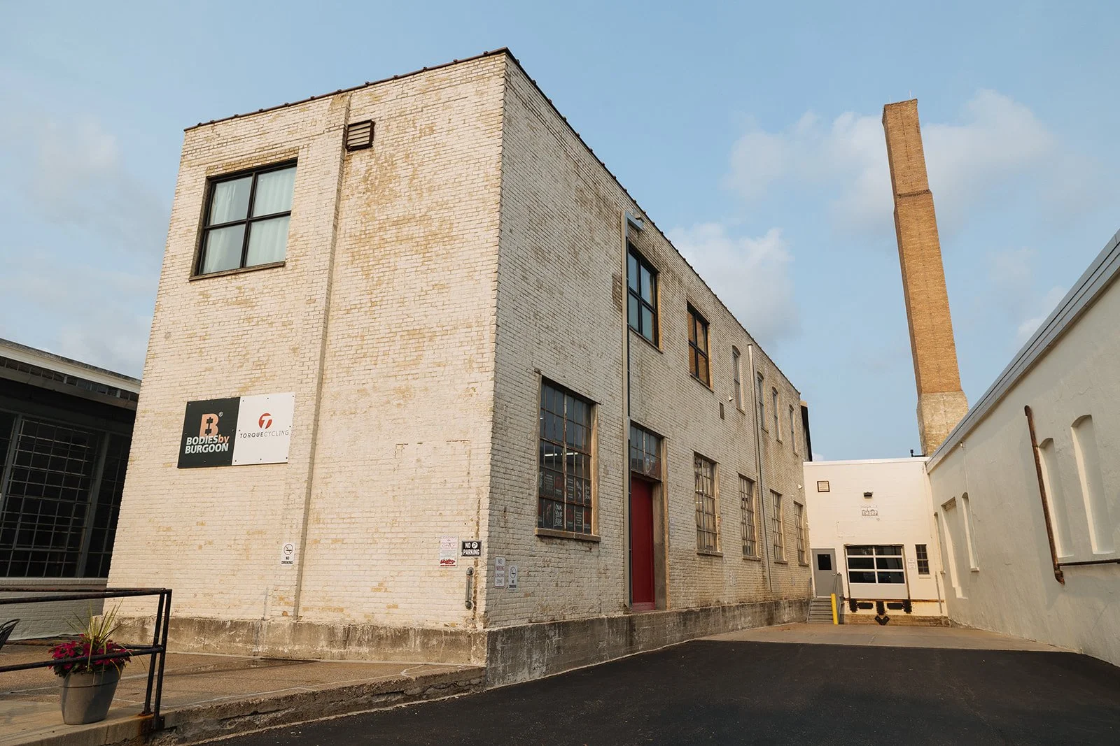 An old industrial brick building with large windows and a red door, set against a blue sky with a tall chimney in the background, and signs on the wall indicating it is associated with Bodies by Burgoon and Torque Cycling.