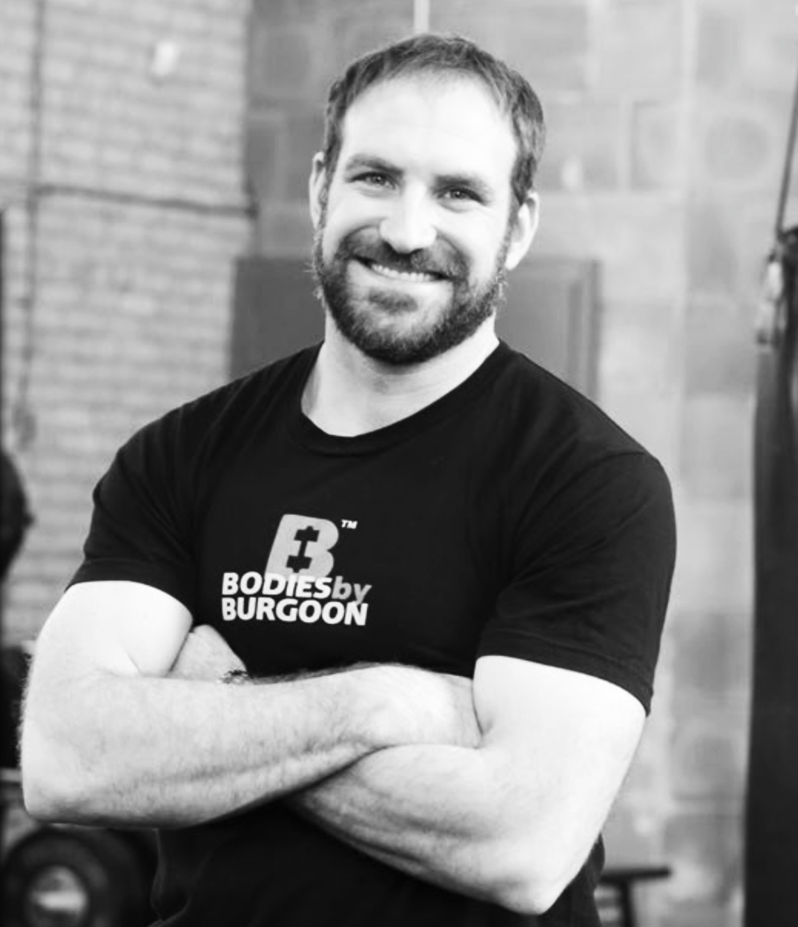 A smiling man with a beard and short hair, crossing his arms, wearing a black T-shirt with 'BODIES by BURGOON' printed on it, standing in a gym with brick walls.