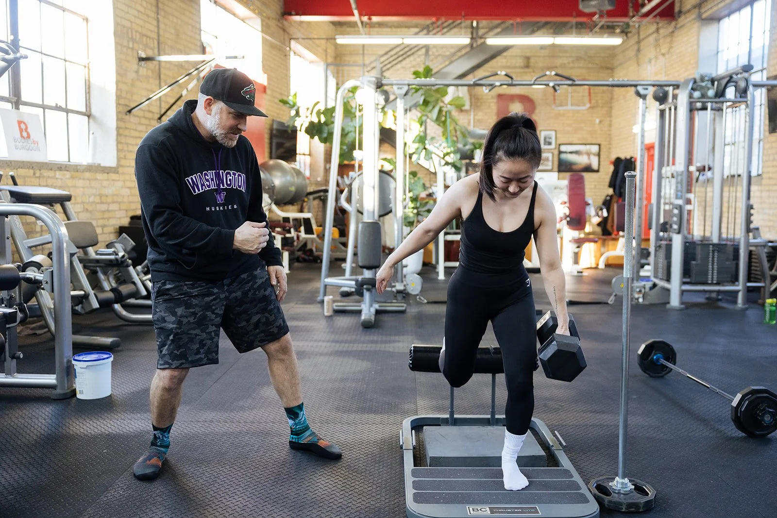 A woman in black workout clothes stepping onto a platform with a dumbbell in each hand while a man in a Washington Huskies hoodie and camouflage shorts coaches her in a gym.