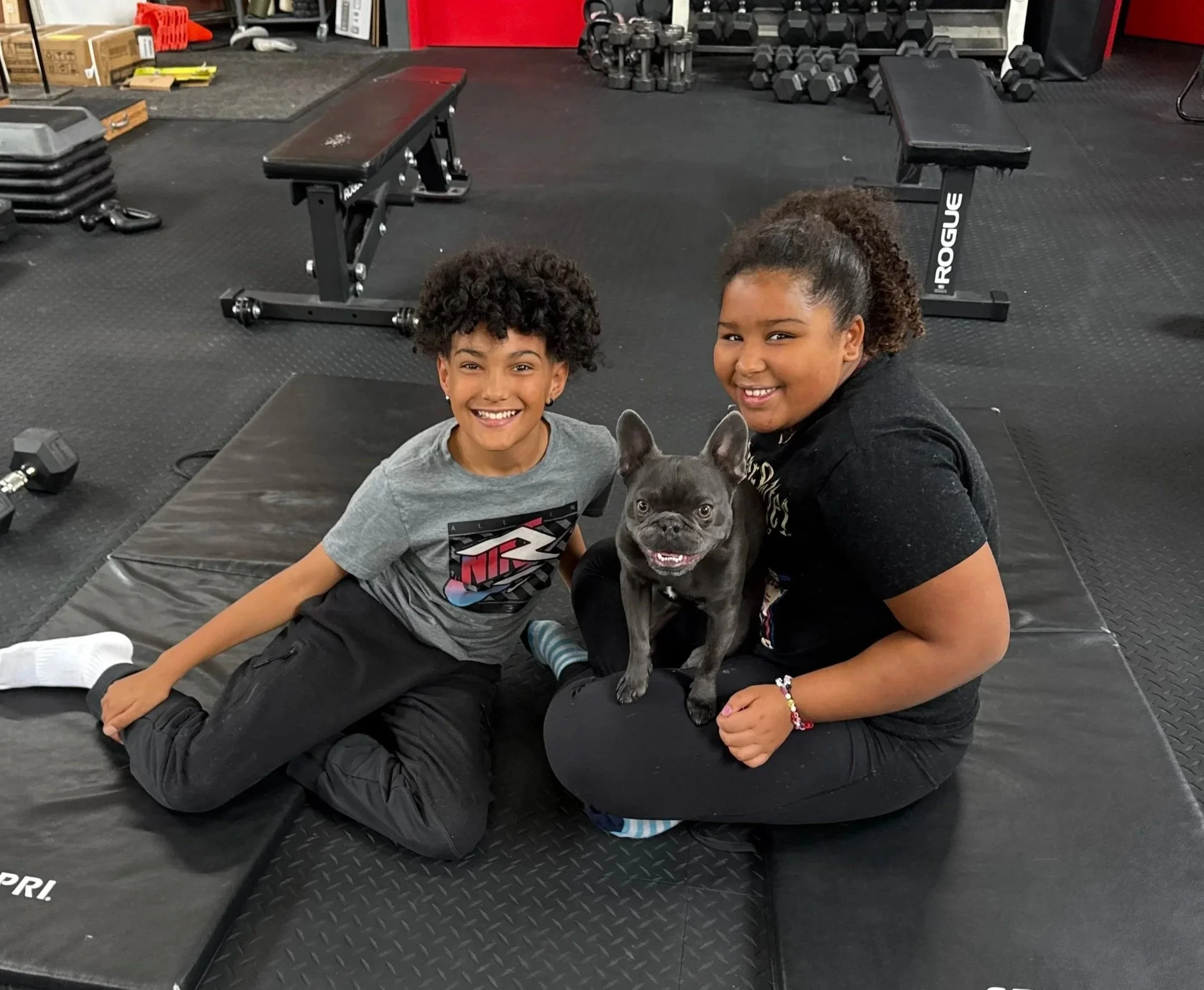 Two kids and a small black French Bulldog sitting on a black exercise mat in a gym, surrounded by gym equipment including dumbbells and benches.