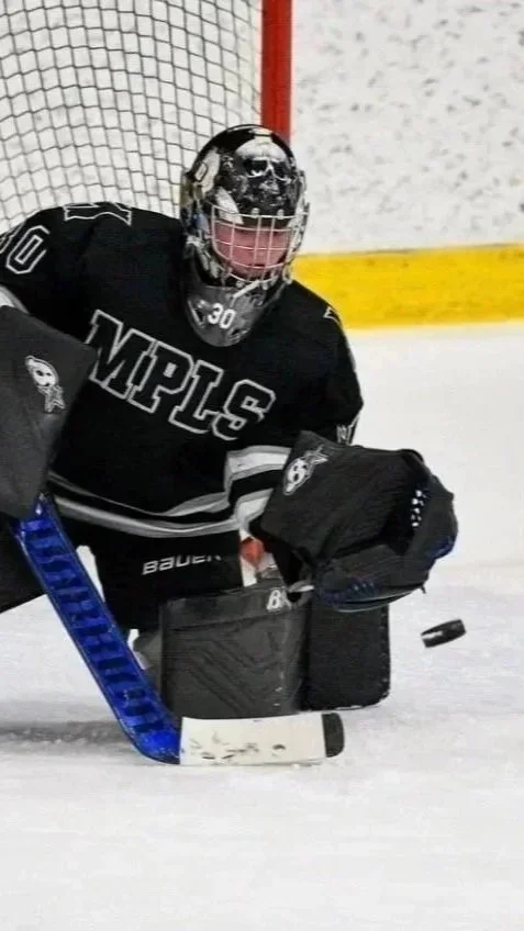 Ice hockey goalie in black jersey with WHITE letters defending near the goal, wearing a mask with a skull design, holding a hockey stick, with a puck on the ice.