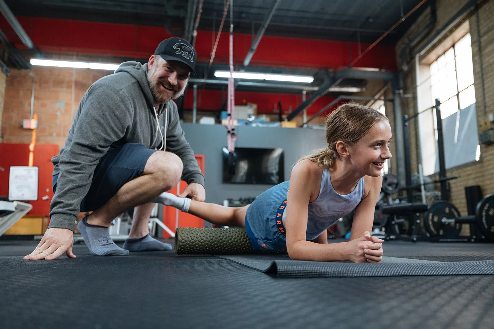 A young girl doing a plank exercise with a foam roller under her legs, on a gym mat, while an adult man supports her by holding her ankles, both smiling inside a gym.