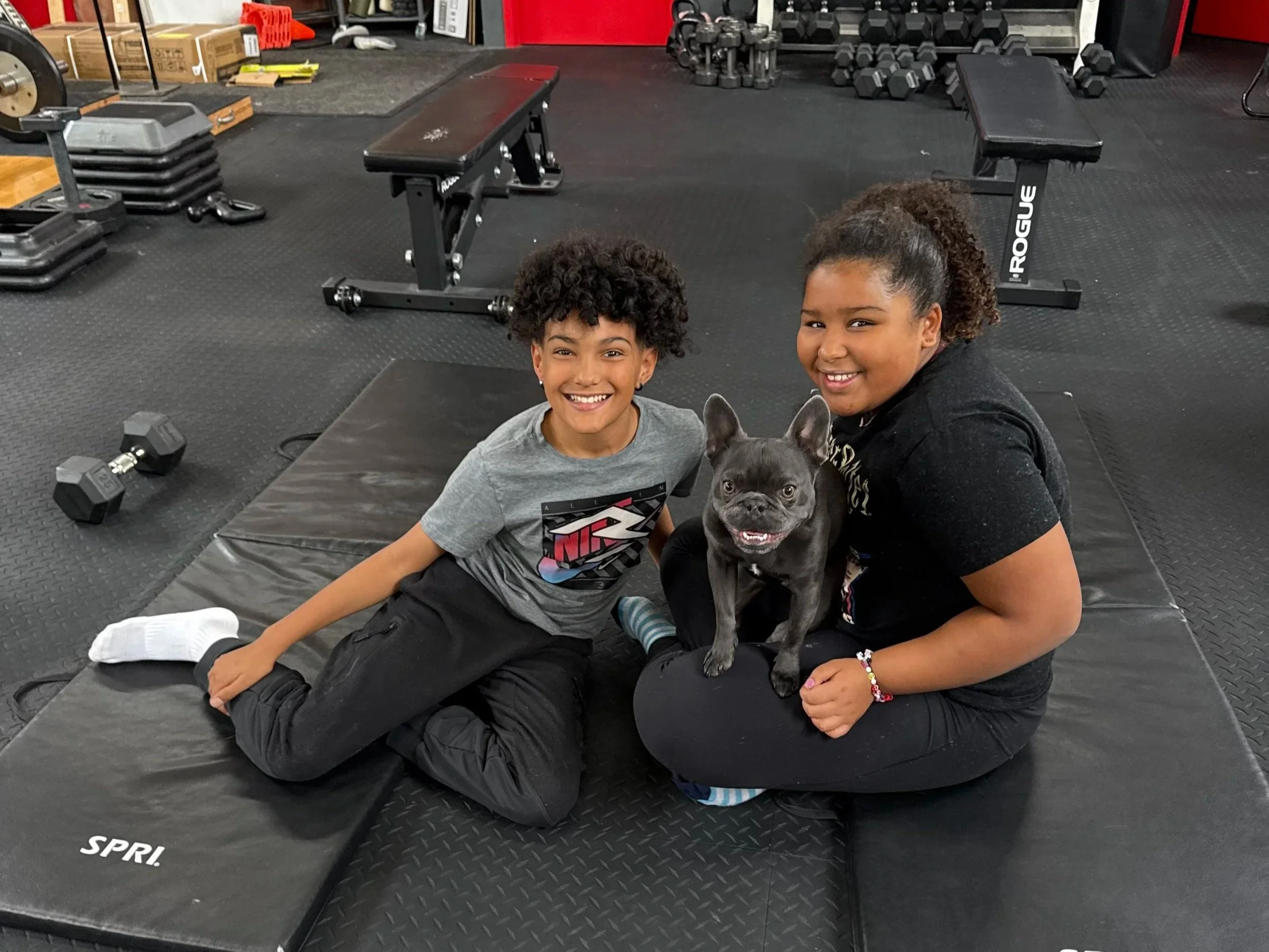 Two smiling kidswith curly hair sitting on gym mats in a gym, holding a French Bulldog dog. The kids are wearing casual athletic clothing. The background has workout equipment like dumbbells, kettlebells, stacked weight plates, and exercise benches.