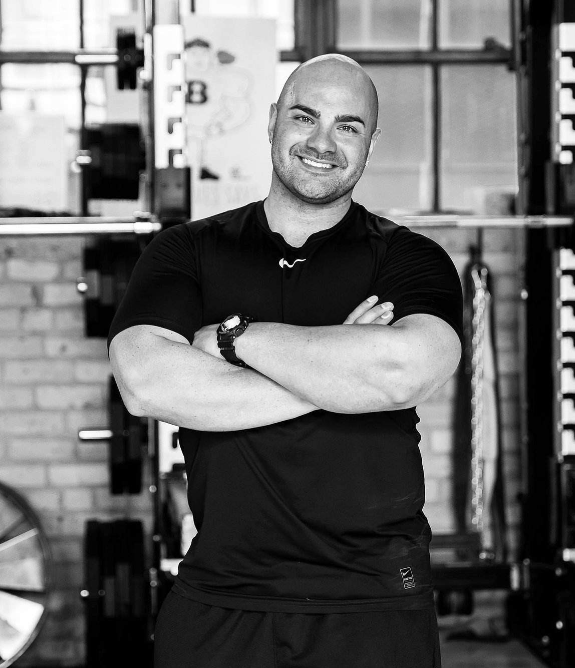 A fit man with arms crossed, smiling, in a gym, wearing a black athletic shirt and watch.
