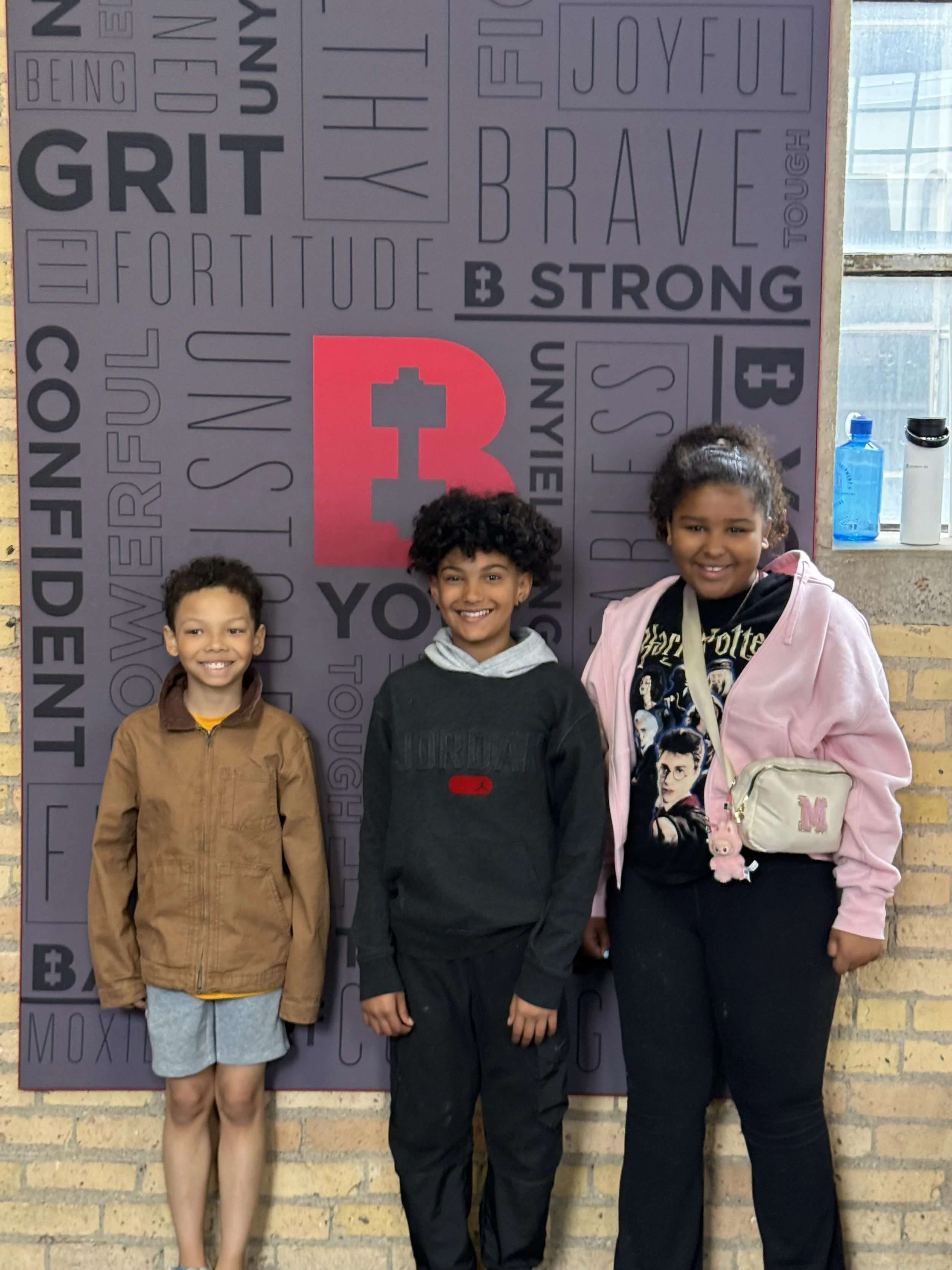 Three children standing in front of a purple wall with positive words and symbols. The children are smiling and looking at the camera. The girl on the right is wearing a white purse with a pink letter 'M' on it, a black Harry Potter shirt, a pink jacket, and has curly hair. The boy in the middle has curly hair, wearing a black hoodie. The girl on the left is wearing a tan jacket, gray shorts, and has straight hair.