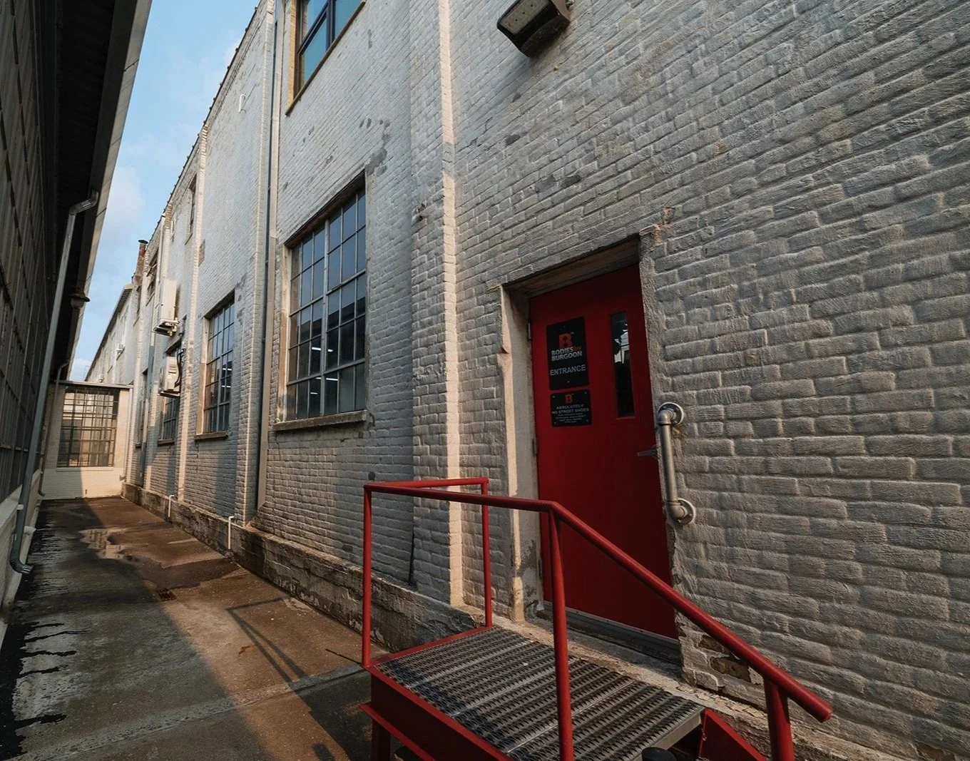 Back alley with brick wall building, red door with signs, metal handrail, industrial style windows, and narrow alleyway with wet pavement.
