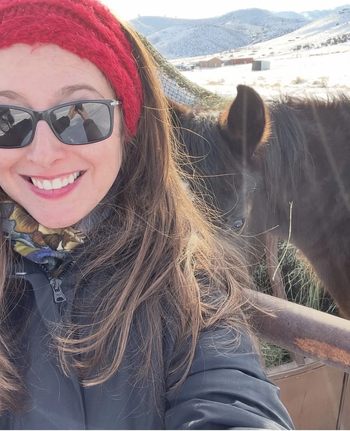 Woman smiling wearing sunglasses and a red knit headband, standing next to a dark horse with a mountain landscape in the background.