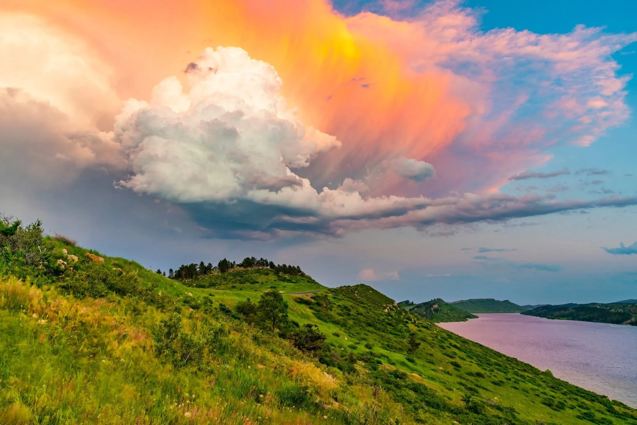 Scenic view of a river flowing beside a lush green hillside under a colorful sky with large clouds and a vibrant sunset.