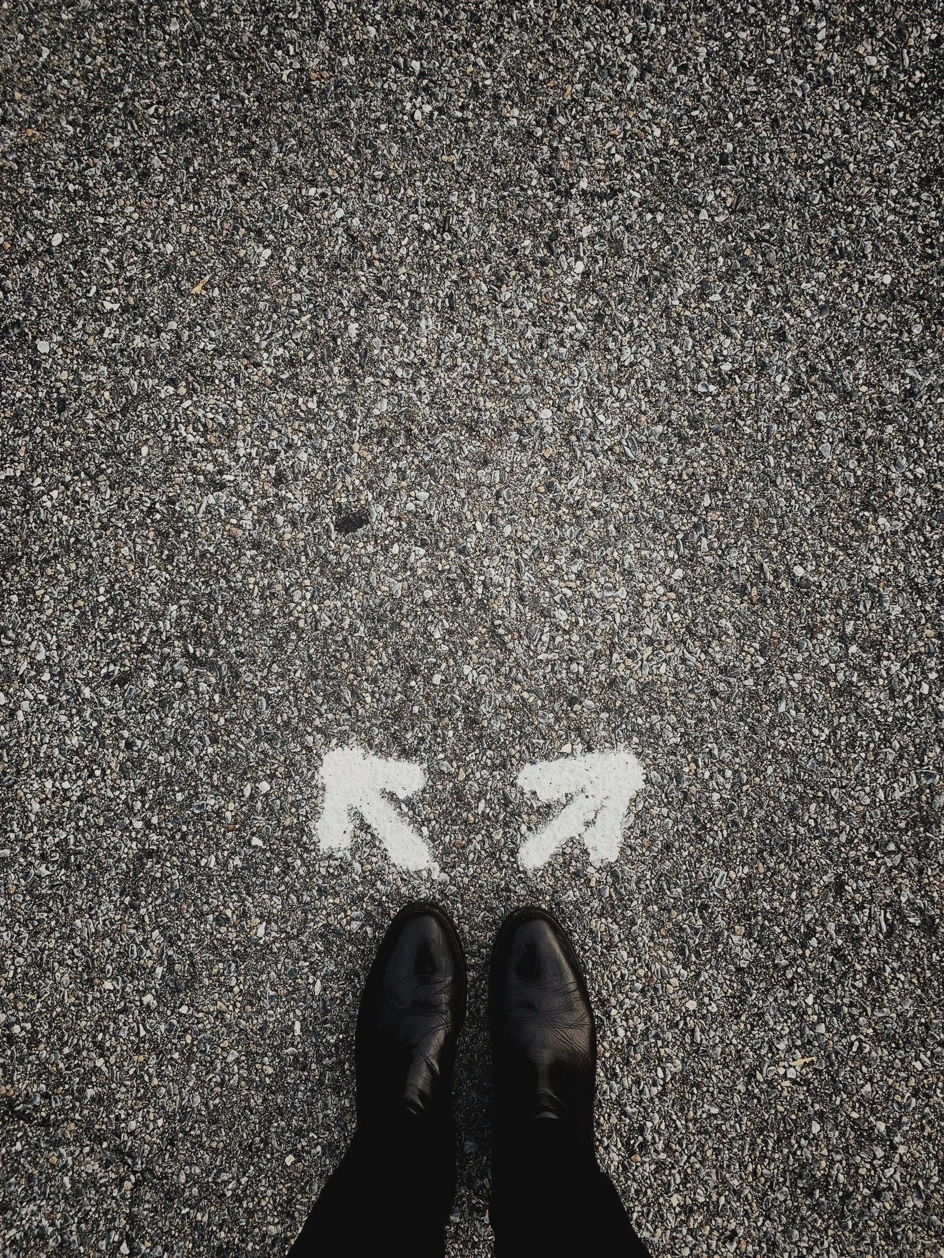 Photo taken from above shows black boots standing on gravel ground with two white arrows painted on the ground pointing in opposite directions.