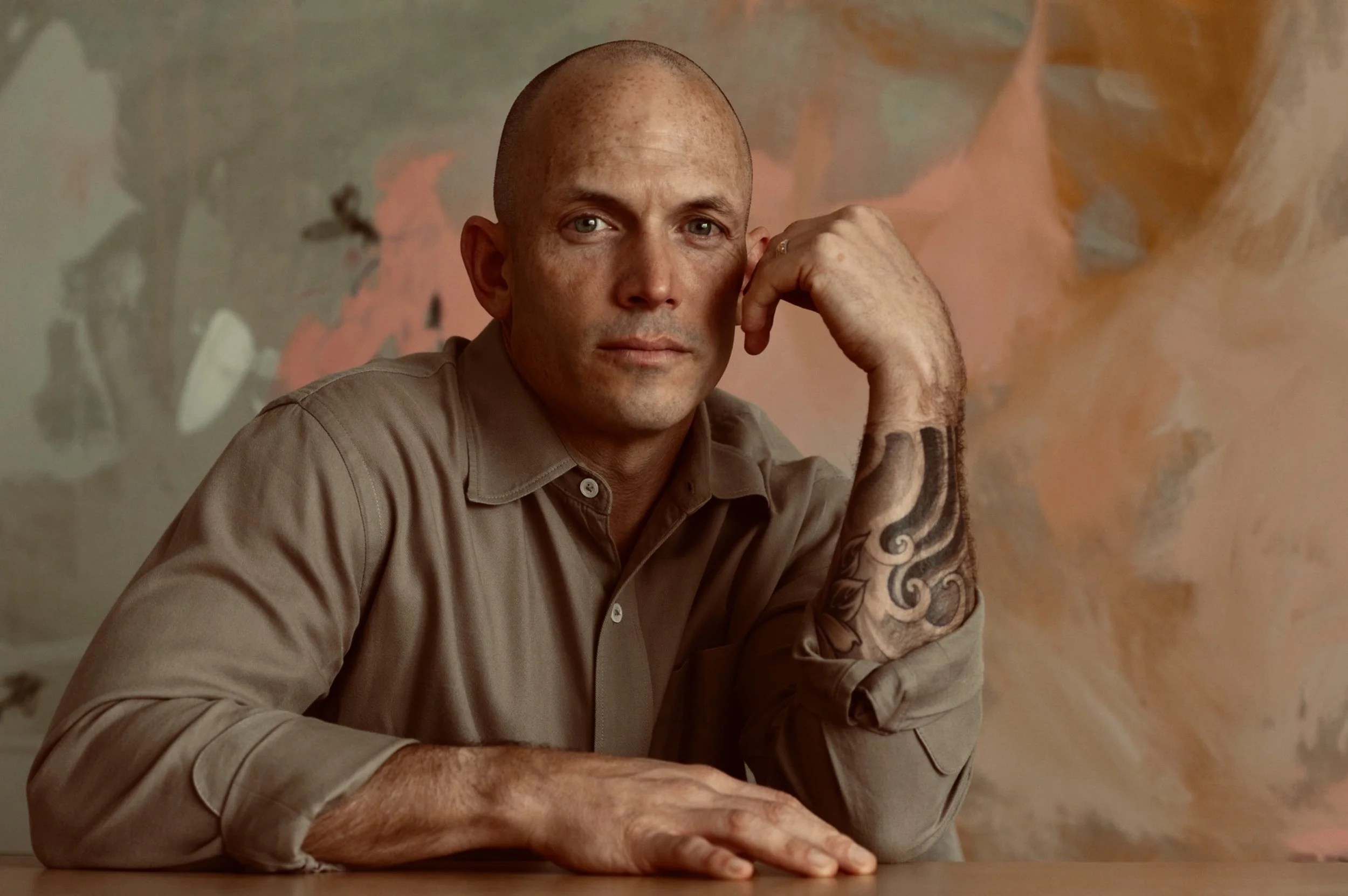 A bald man with a tattoo on his right arm, wearing a beige shirt, sitting in front of a colorful abstract background.