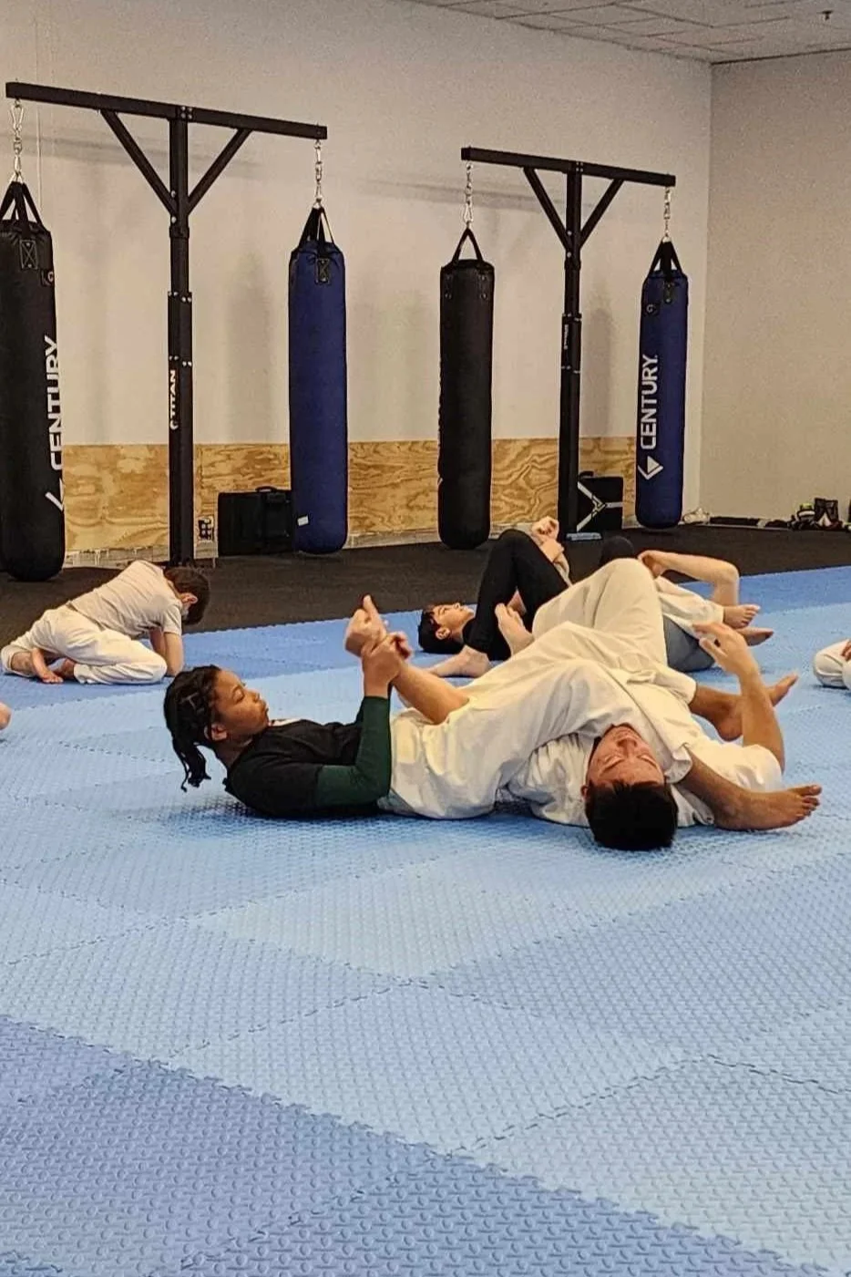 Martial arts class practicing grappling and submission techniques on blue mats in a gym with punching bags in the background.