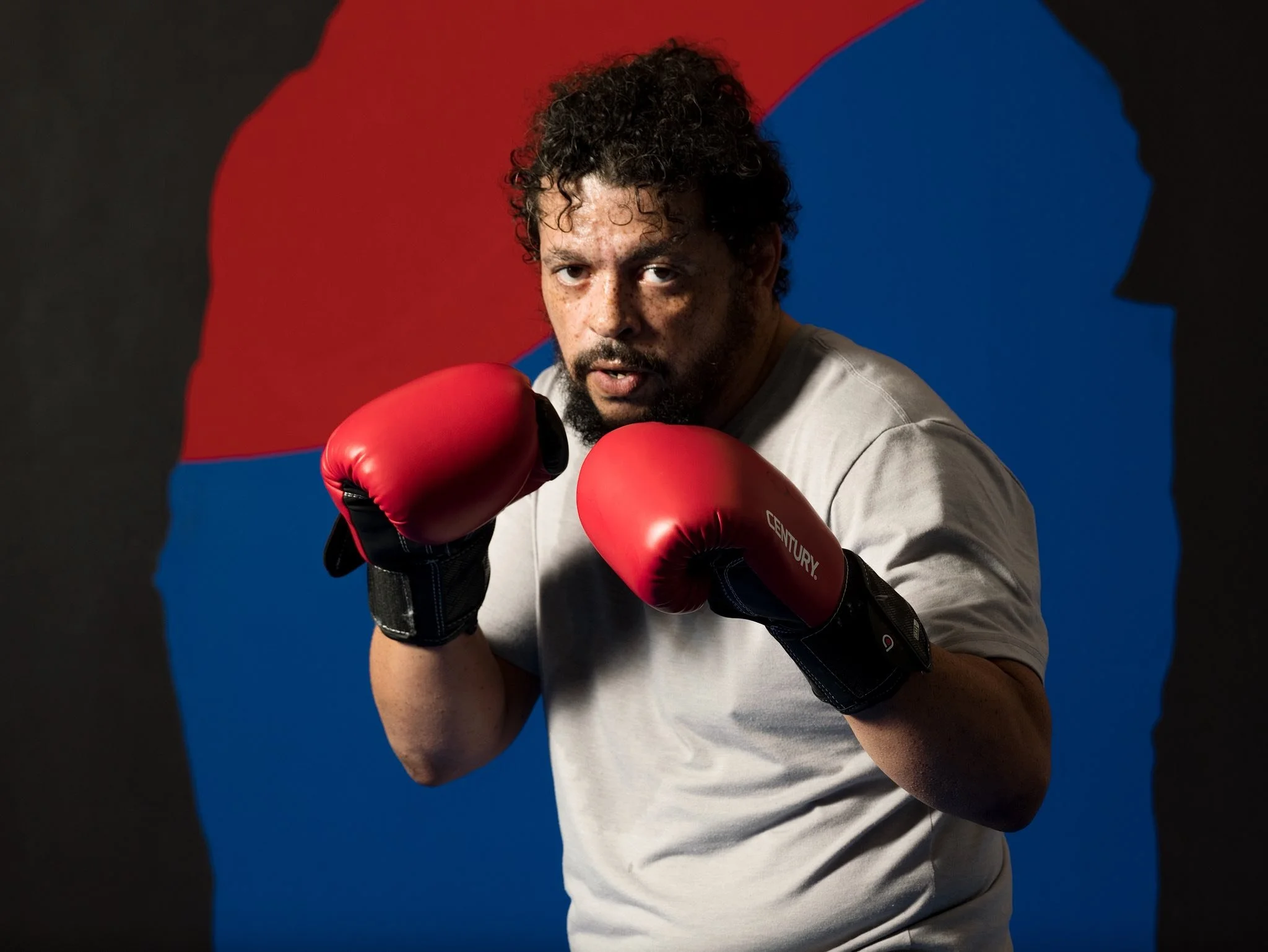 A man with curly hair and a beard wearing red boxing gloves and a light-colored t-shirt, standing in a fighting stance against a background of a silhouette of two heads, one red and one blue.