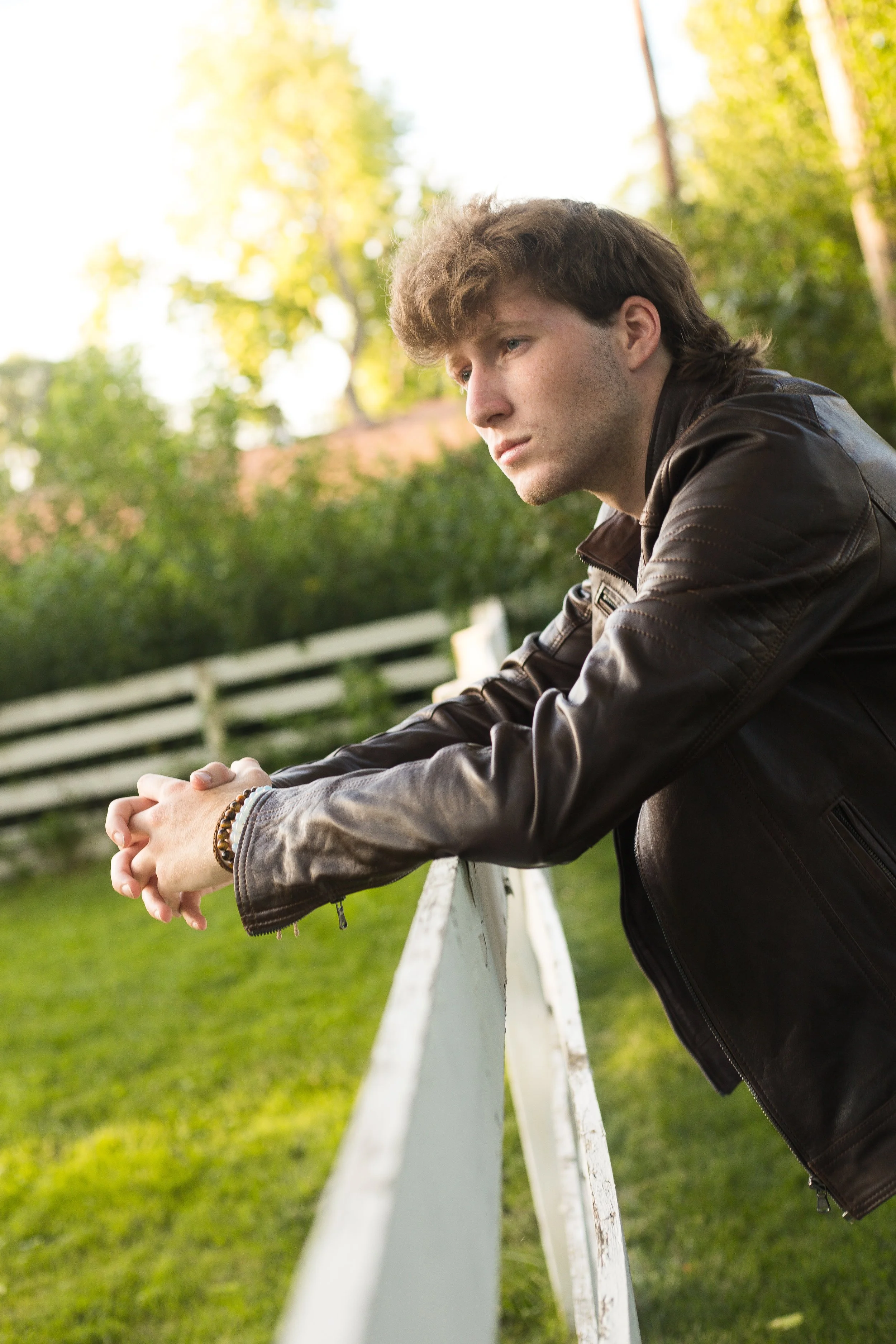 A young man in a dark leather jacket leaning on a white fence outdoors, with a contemplative expression and green trees in the background.