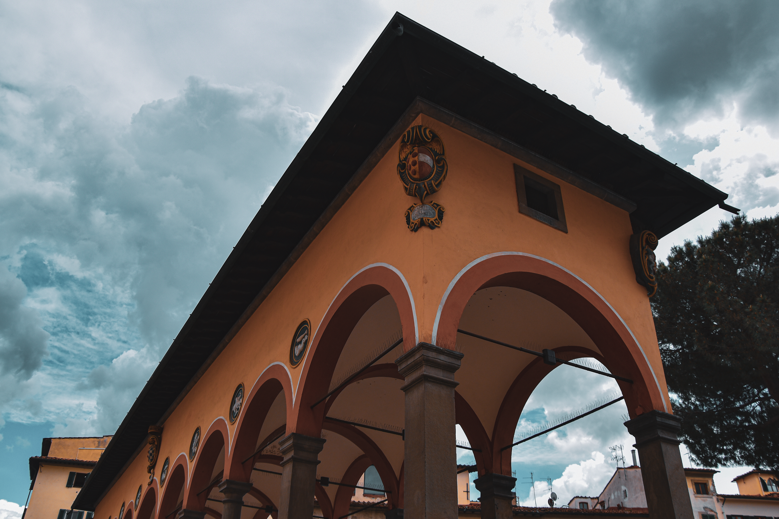 A building with orange walls and arched openings, decorative emblems, and a dark roof, under a cloudy sky.