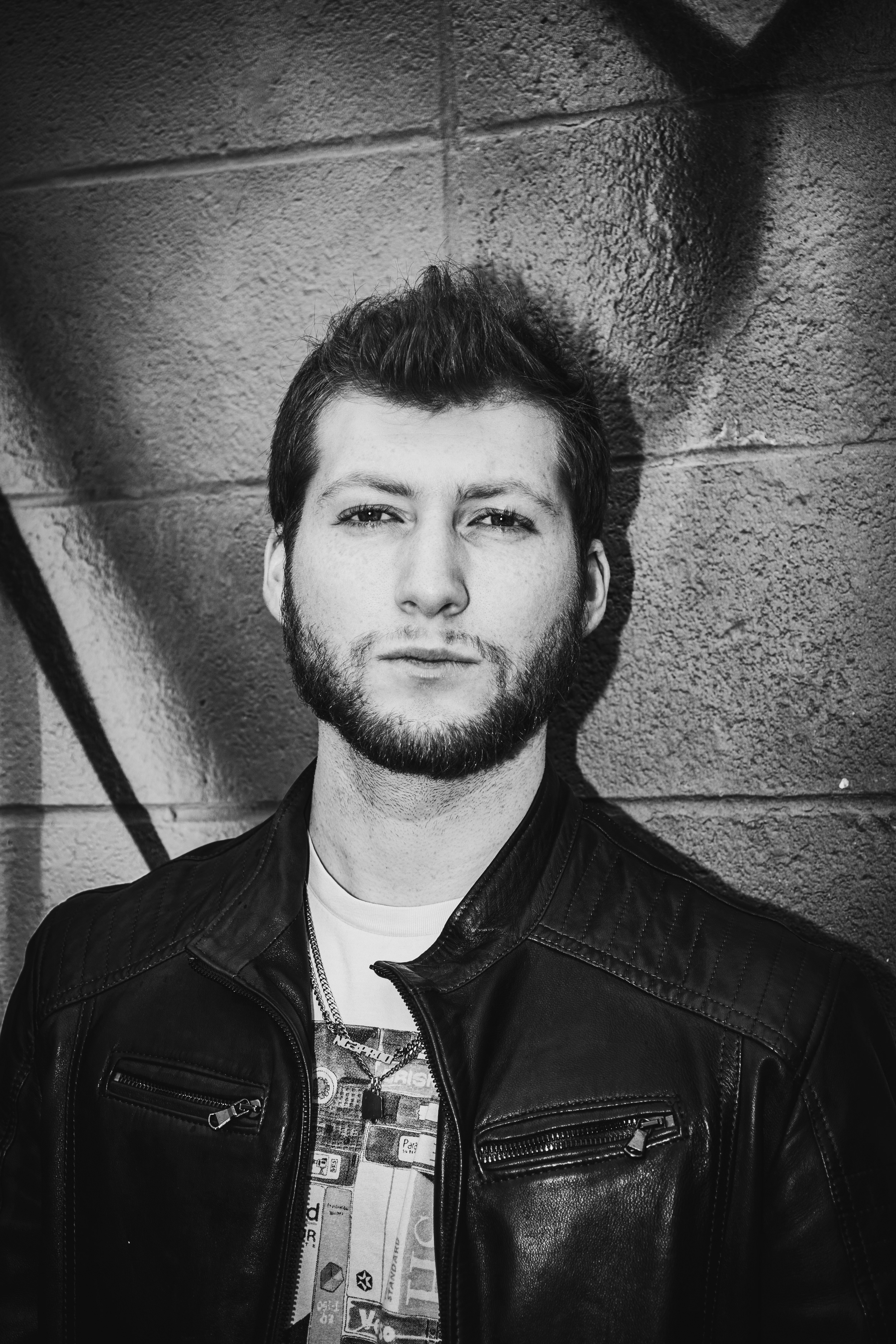 Black and white portrait of a man with a beard and short hair, wearing a leather jacket and a graphic t-shirt, standing against a textured brick wall.