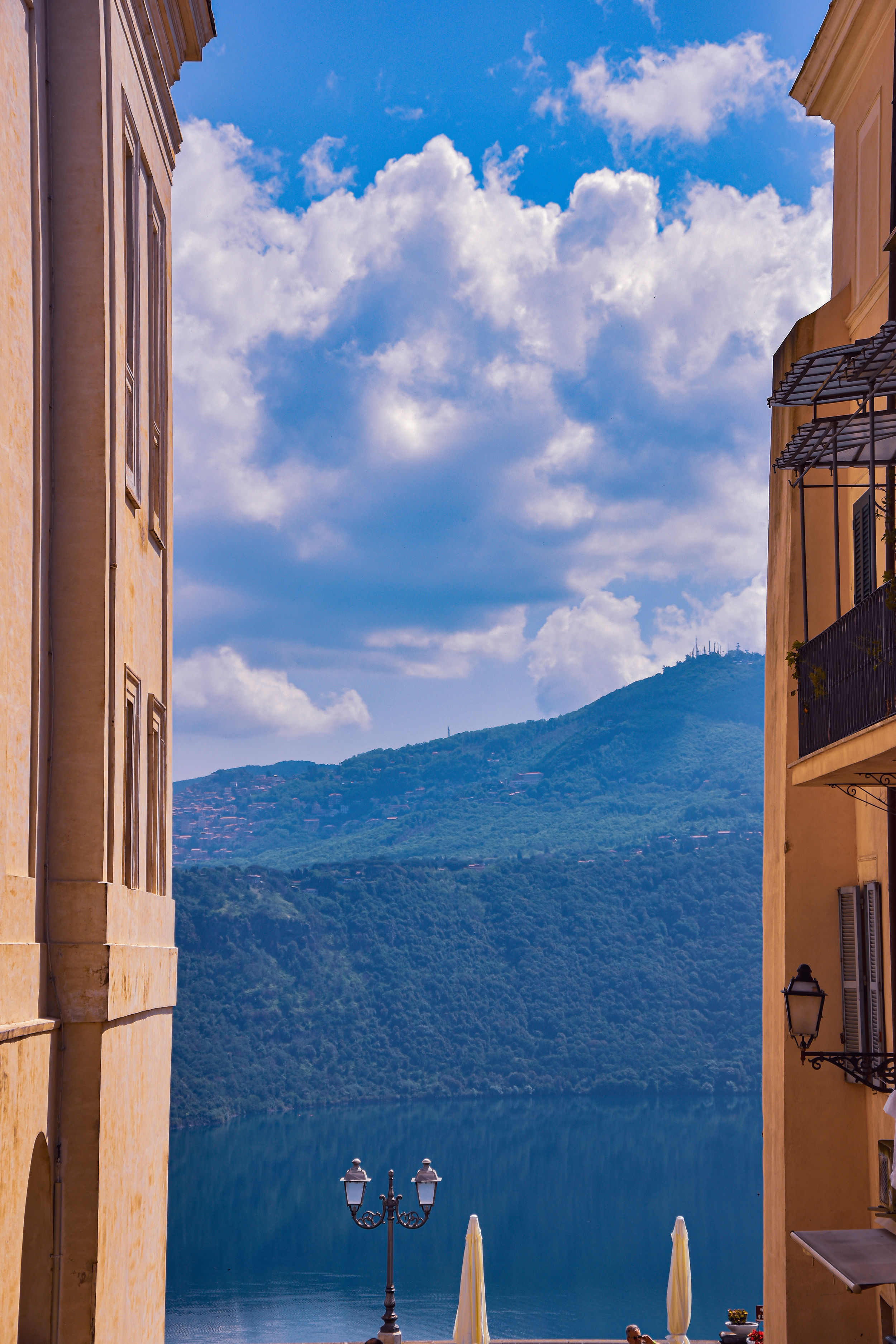 View through a narrow street between two yellowish buildings towards a lake with mountains in the background under a partly cloudy sky.