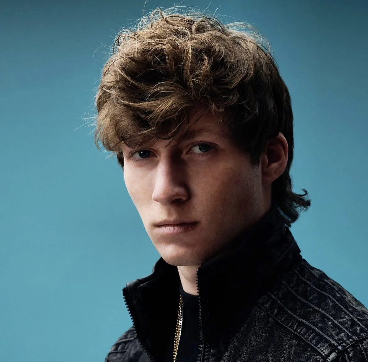 Close-up portrait of a young man with tousled brown hair, wearing a black leather jacket, against a blue background.