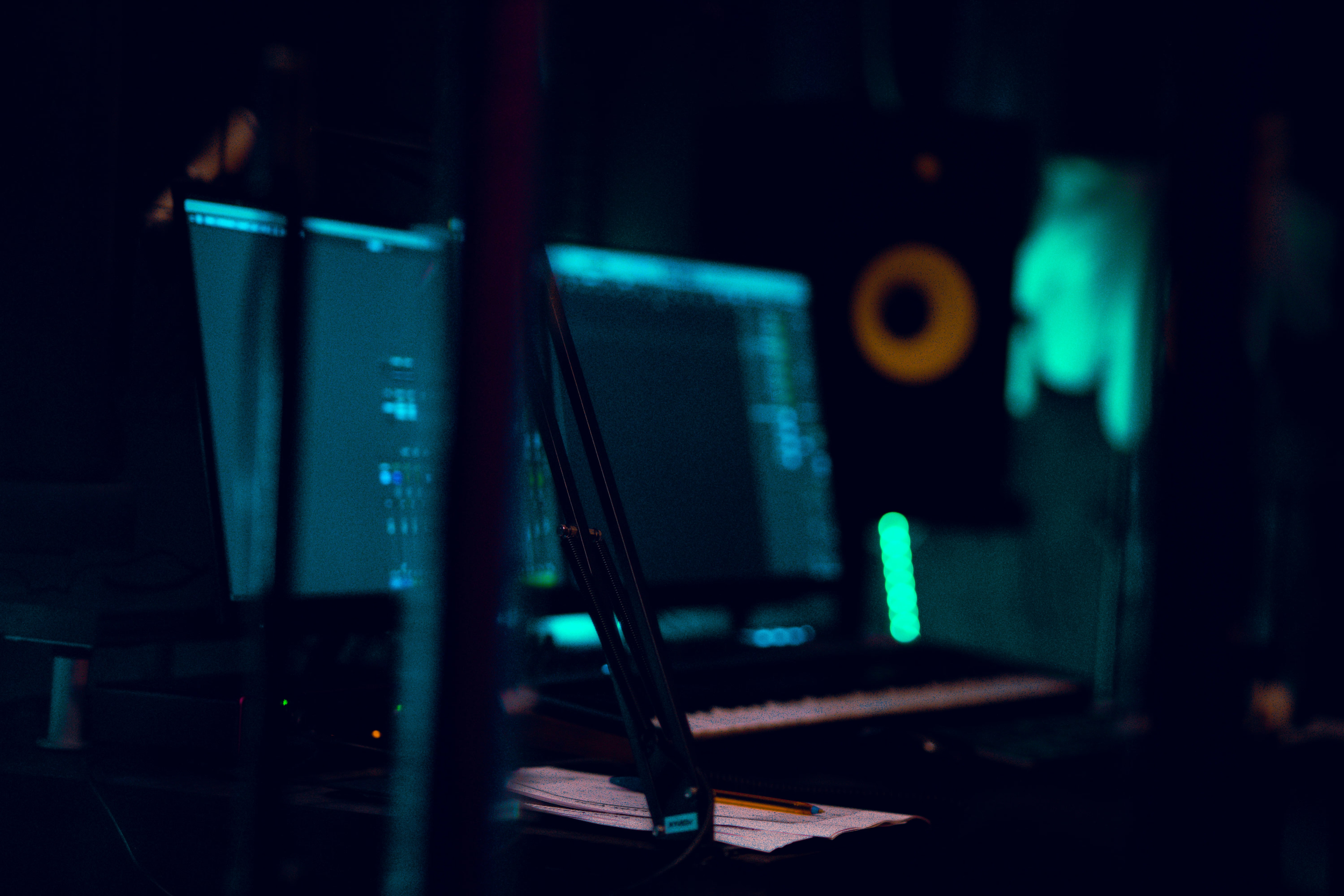 Dimly lit room with computer monitors displaying code, a keyboard, and notes with a pencil on a desk, suggesting a workspace or studio for music production or digital work.