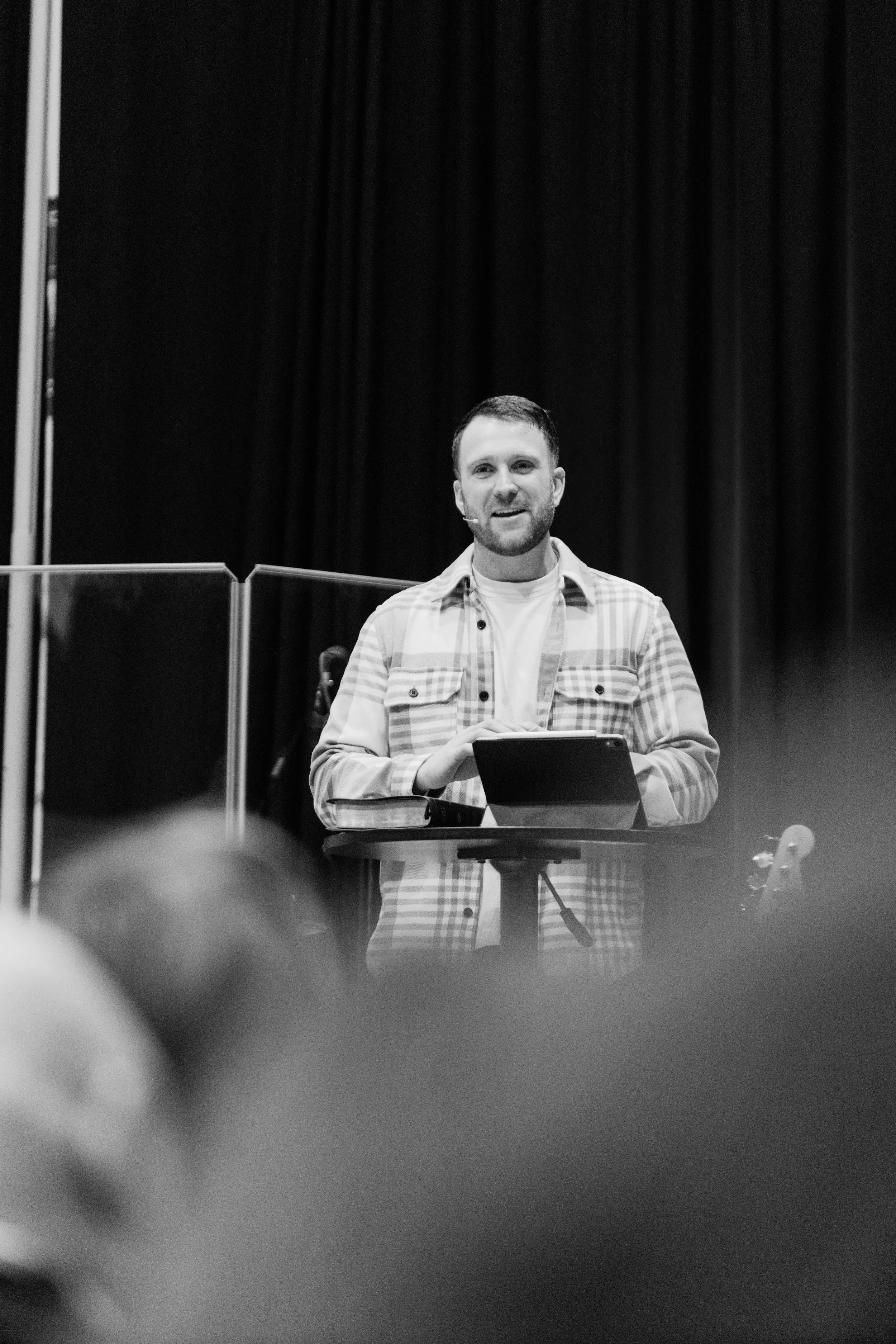 A man standing behind a podium on a stage, speaking into a microphone, with a dark curtain background.