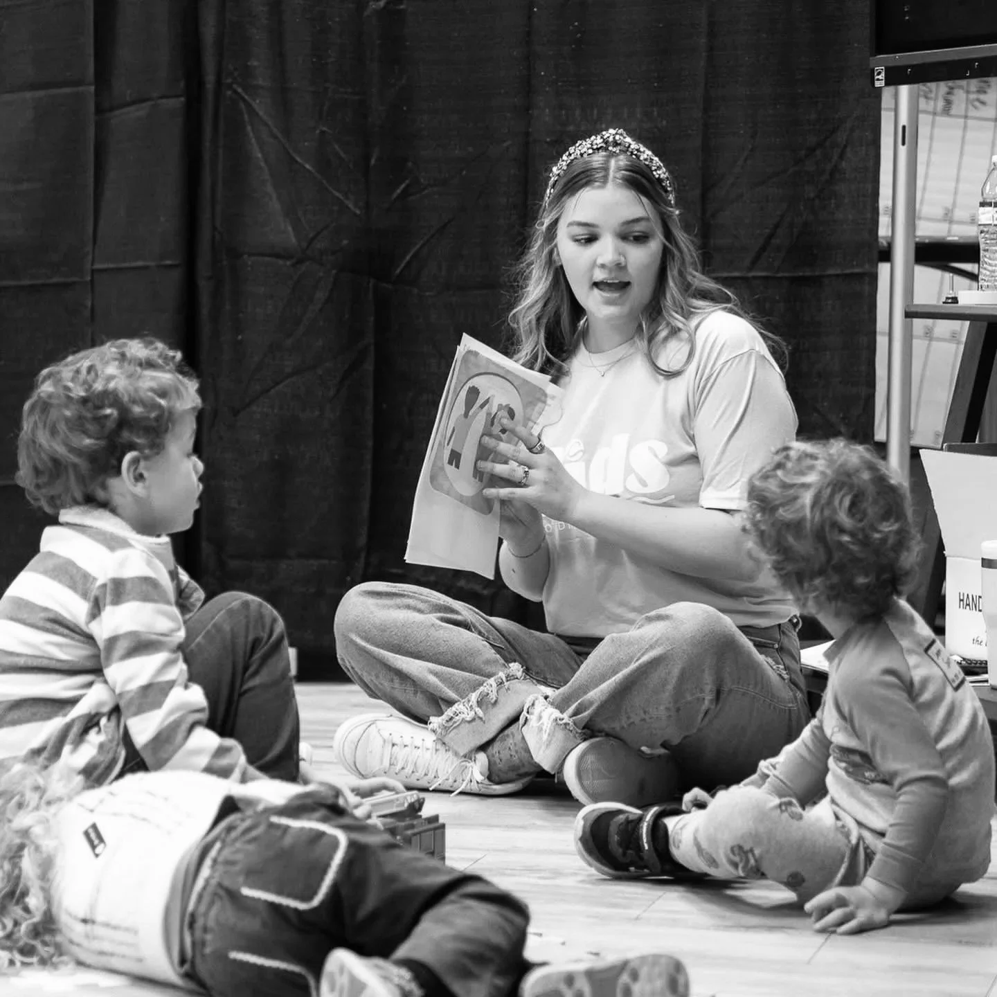 A young woman with a tiara reading a book to children sitting on the floor in a room.