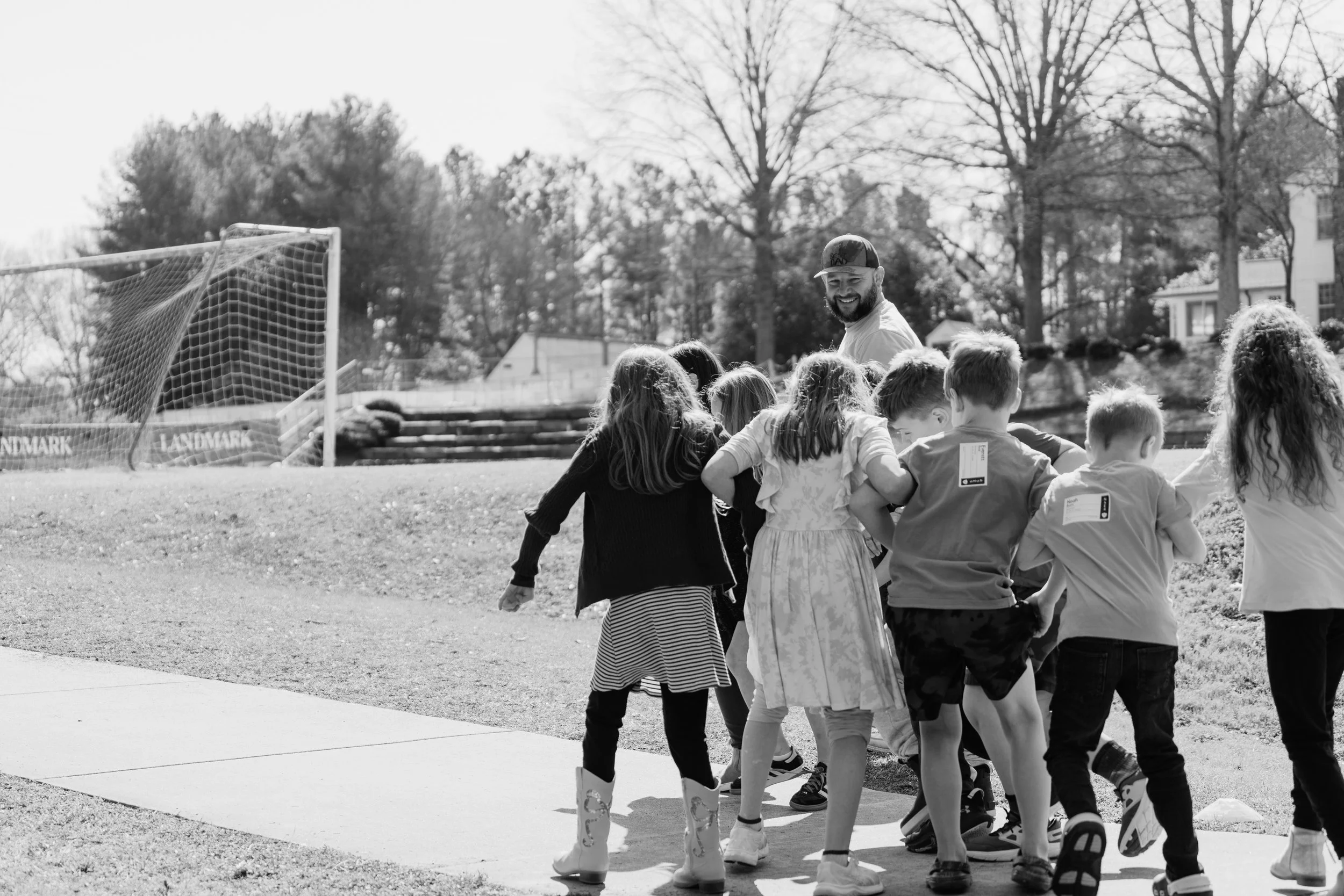 A group of children and a smiling adult man standing outdoors near a soccer goal, with trees and houses in the background.