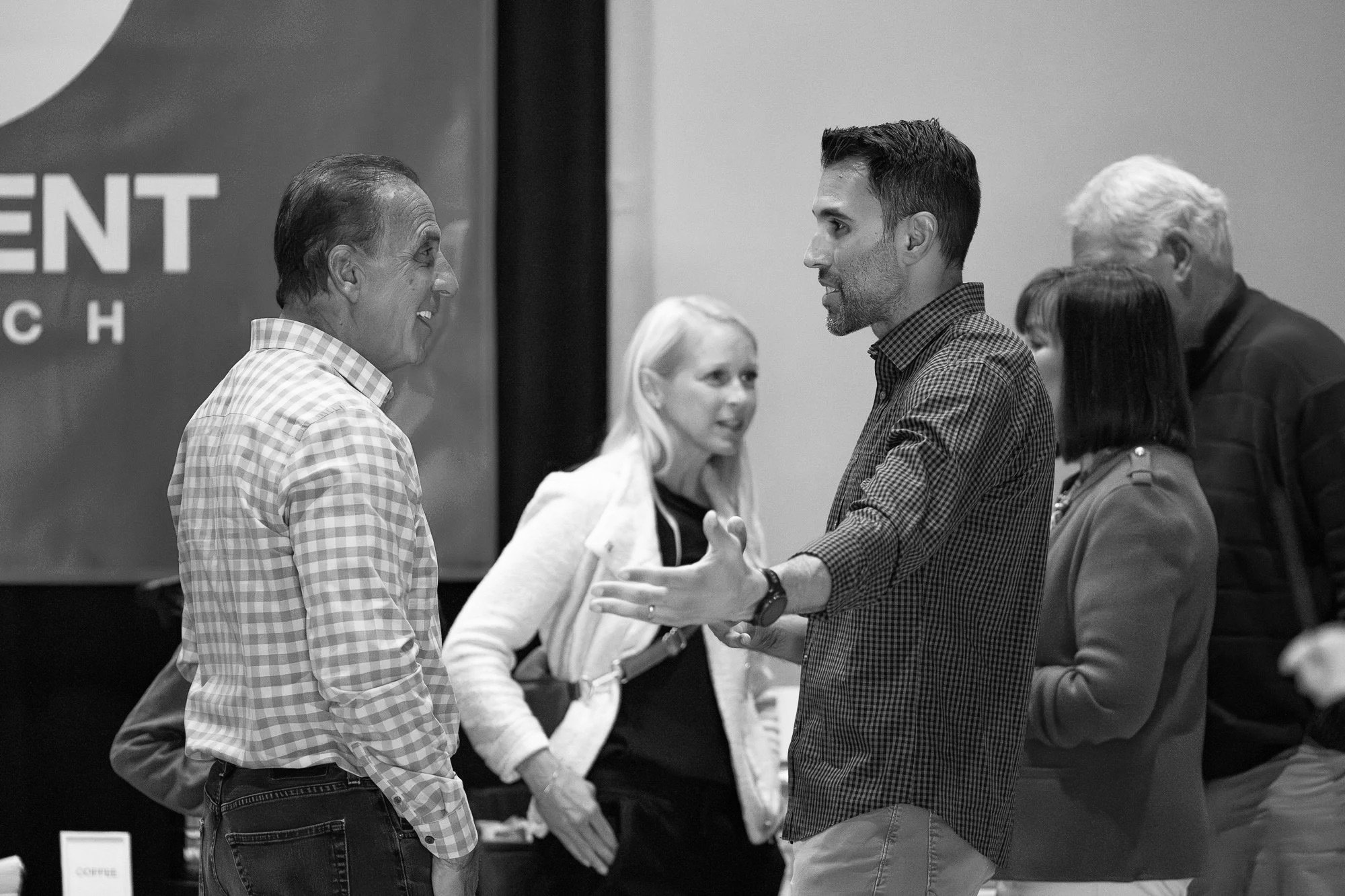 Group of six people engaged in conversation at an indoor event, some smiling and listening.