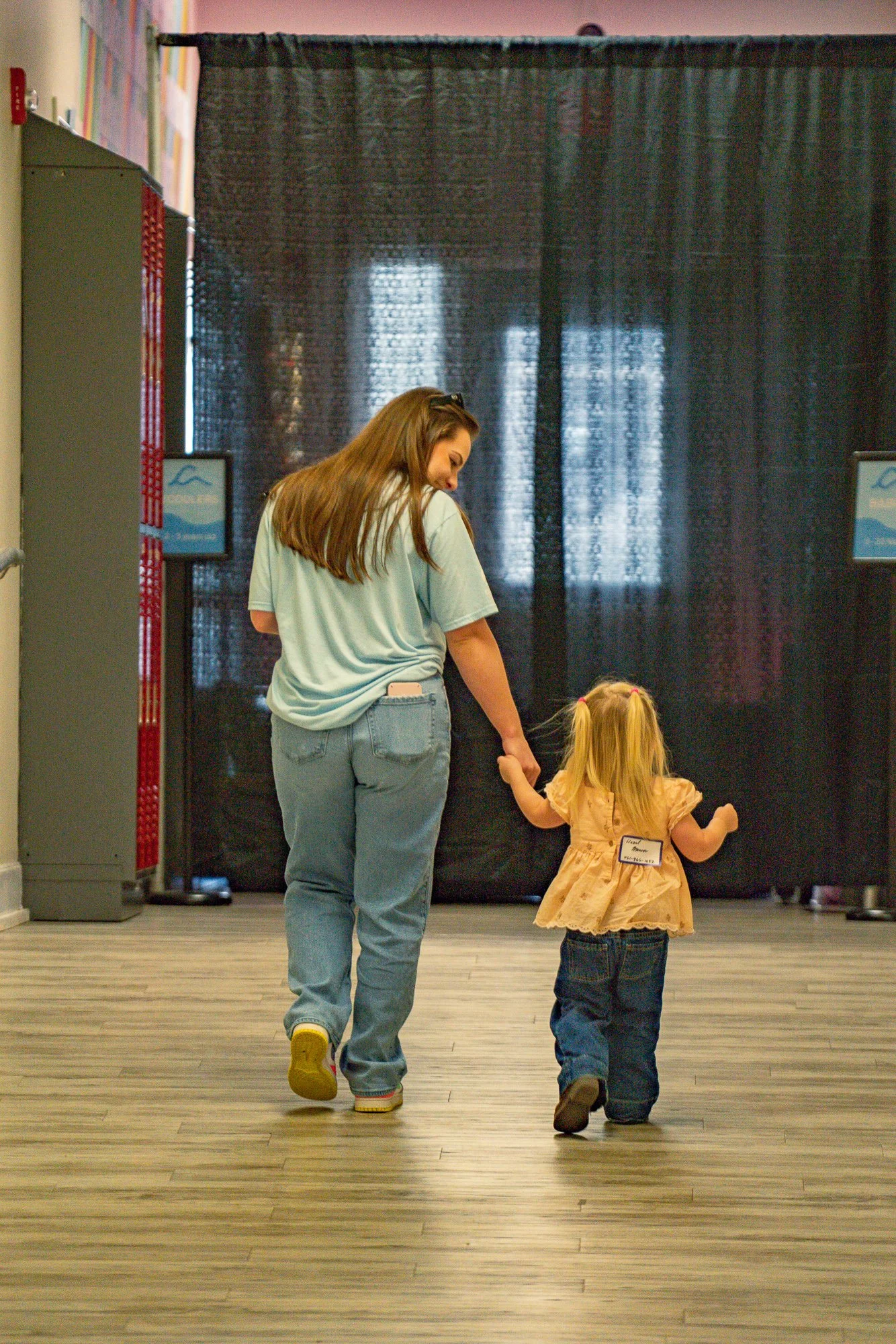 A woman and a young girl holding hands while walking in an indoor space with a black curtain background, wooden floor, and room dividers.
