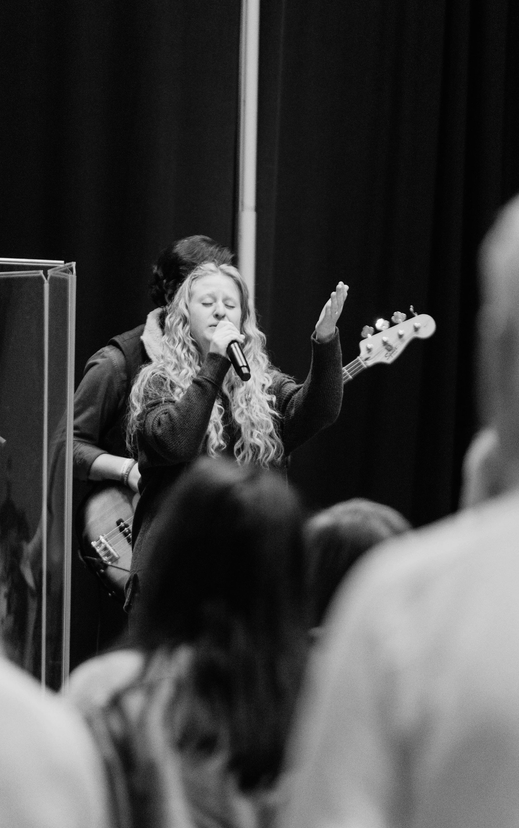 A woman singing into a microphone with eyes closed, surrounded by people, in a black-and-white photo.