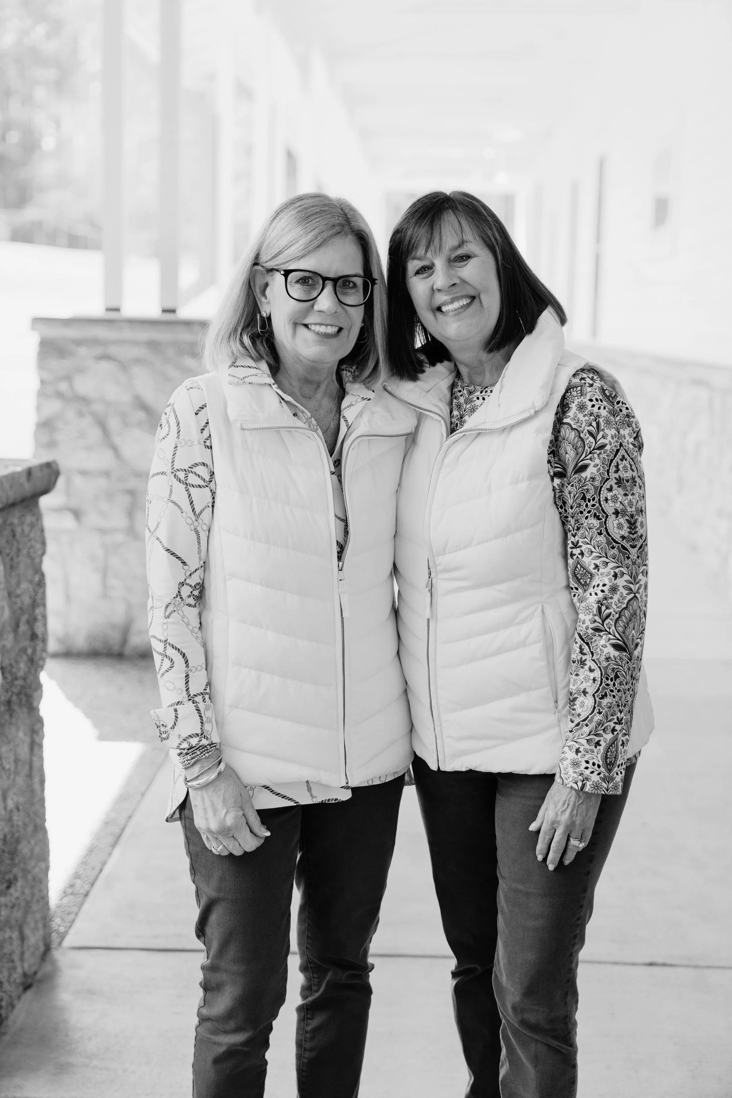 Two women smiling and standing close together outdoors, wearing light-colored puffer vests and patterned long-sleeve shirts in a bright, sunny setting.