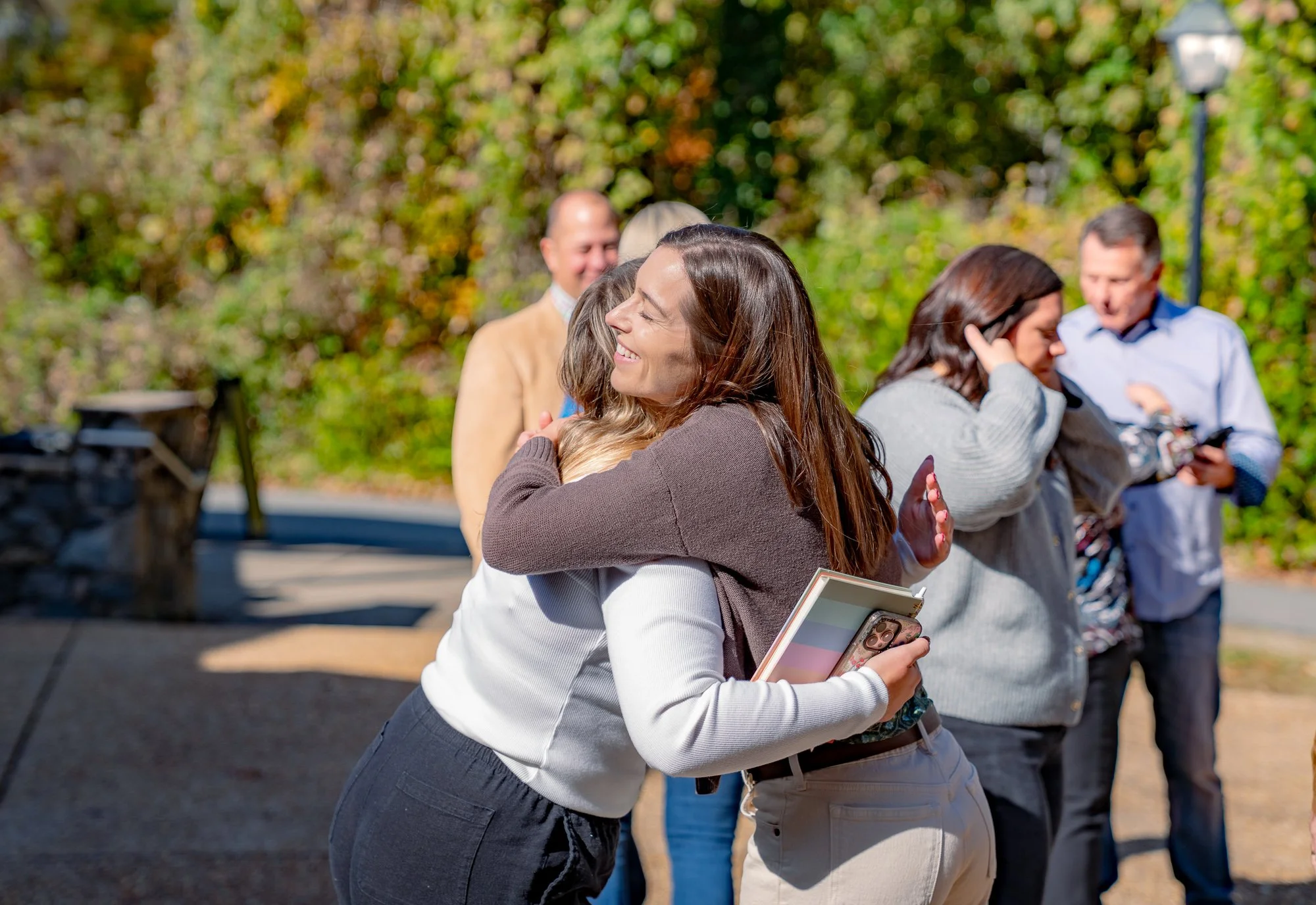 Two women hugging outdoors during daytime with other people in the background, smiling and chatting, surrounded by trees with autumn foliage.