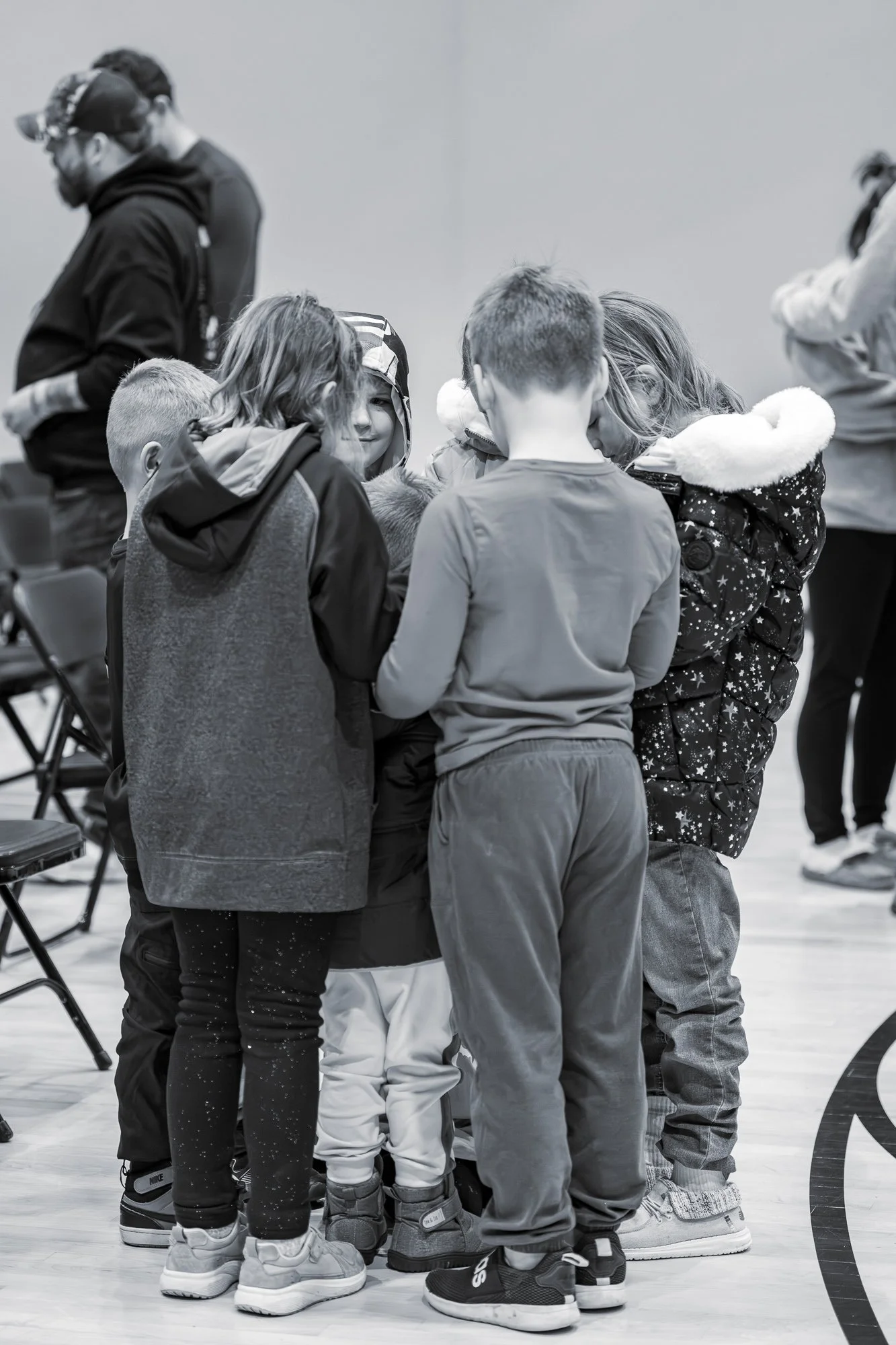 Group of children huddled together in the center, examining or playing with something, with two adults standing in the background, in a gymnasium or indoor sports facility.