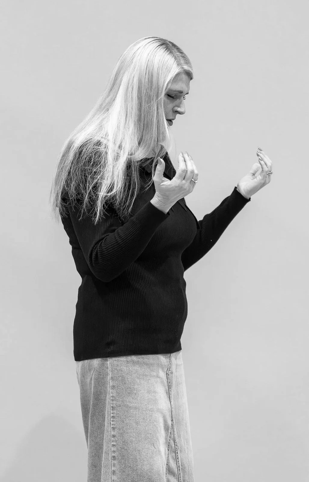 A woman with long blonde hair, wearing a black sweater and light-colored jeans, looking down with her hands raised near her chest in front of a plain wall.